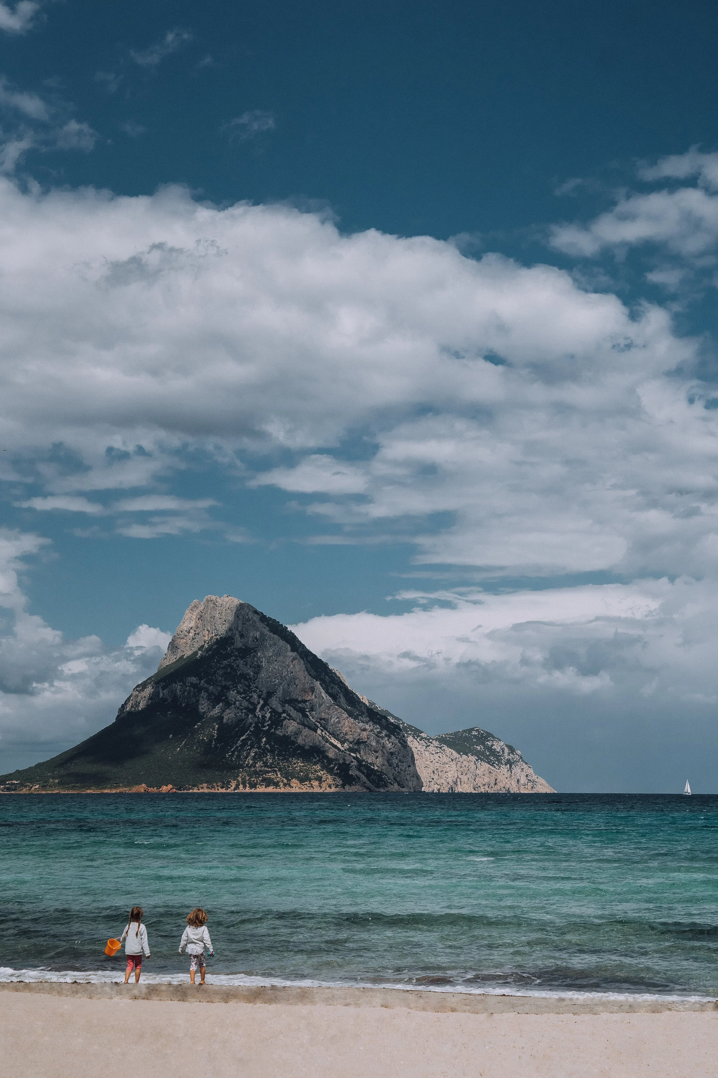 Two children on a beach facing the ocean with a mountain in the background and a sailboat on the water.