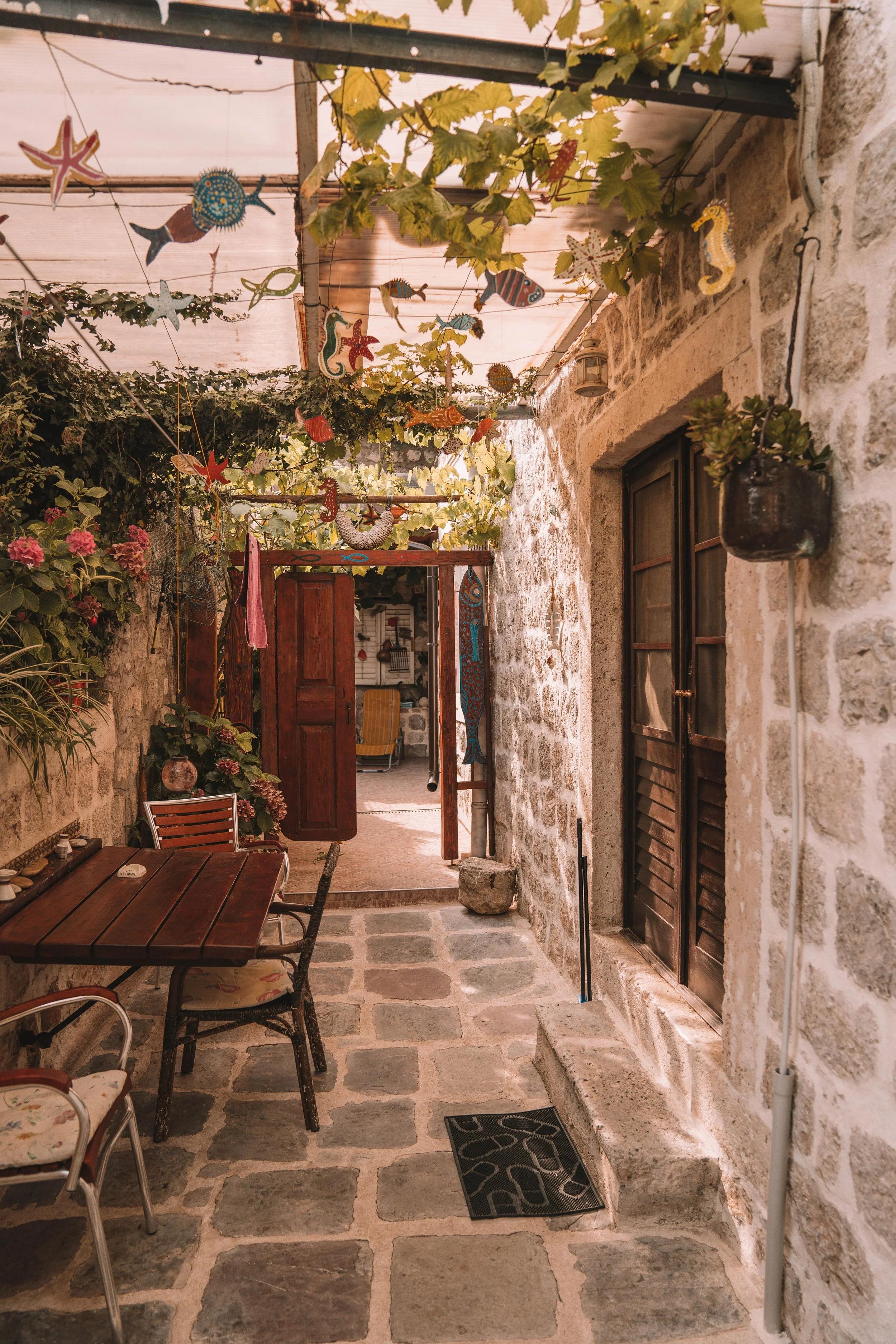 Stone-paved courtyard with a wooden table and chairs, surrounded by stone walls and decorated with hanging sea-themed ornaments and plants.