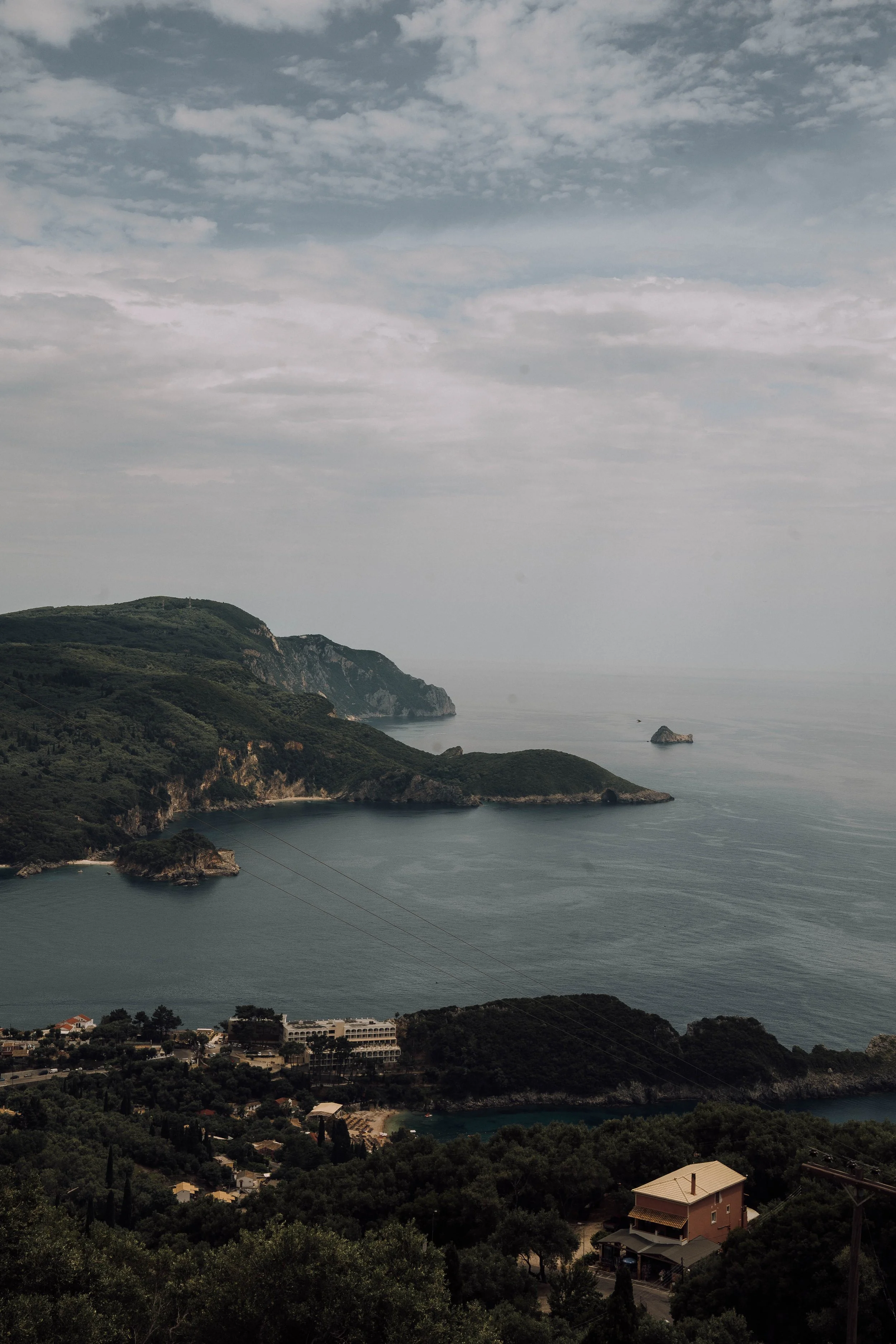 Coastal landscape with green hills, ocean, and scattered buildings near the shoreline under a partly cloudy sky.