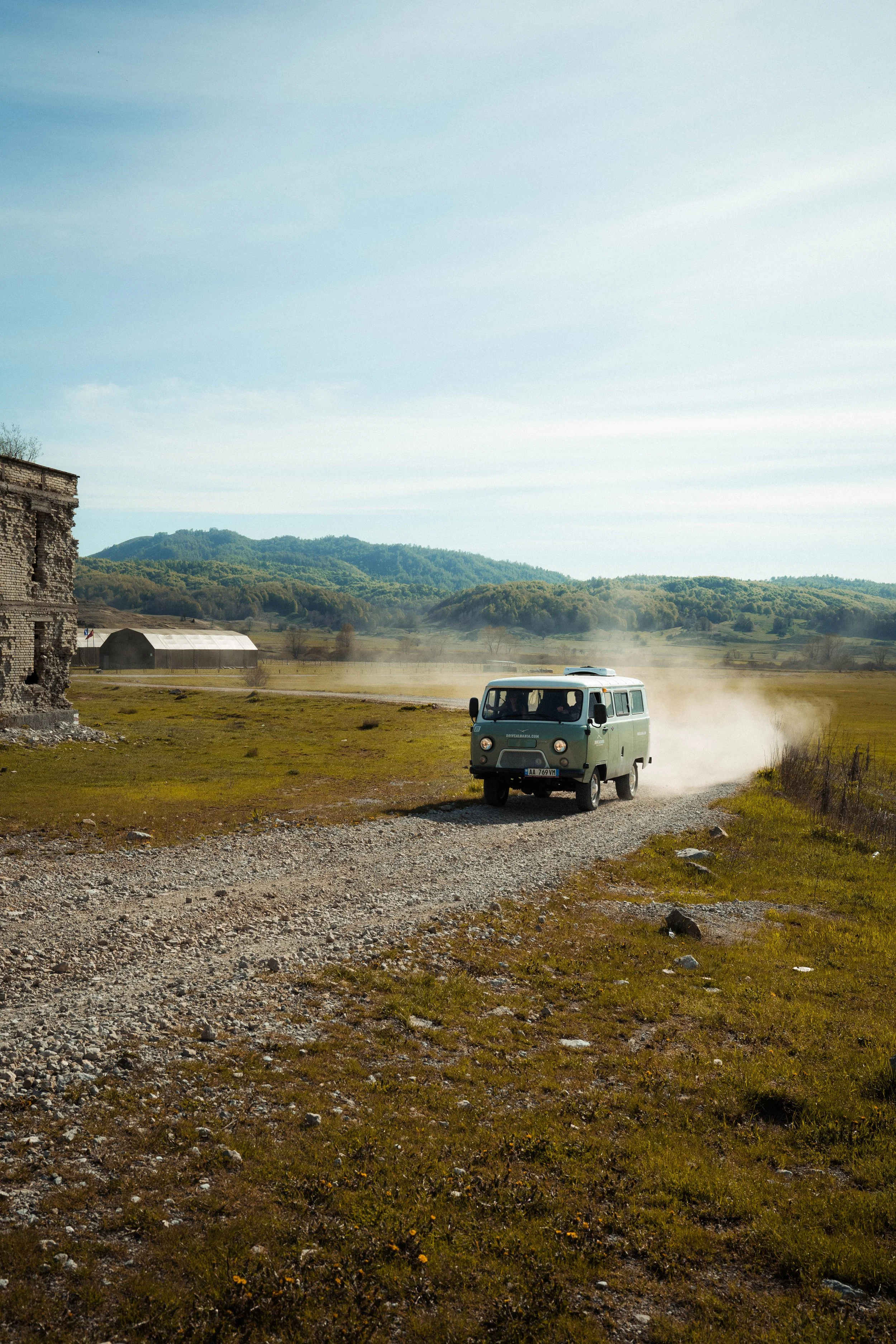 Vintage van driving on a gravel road in a rural landscape with mountains and blue sky in the background.