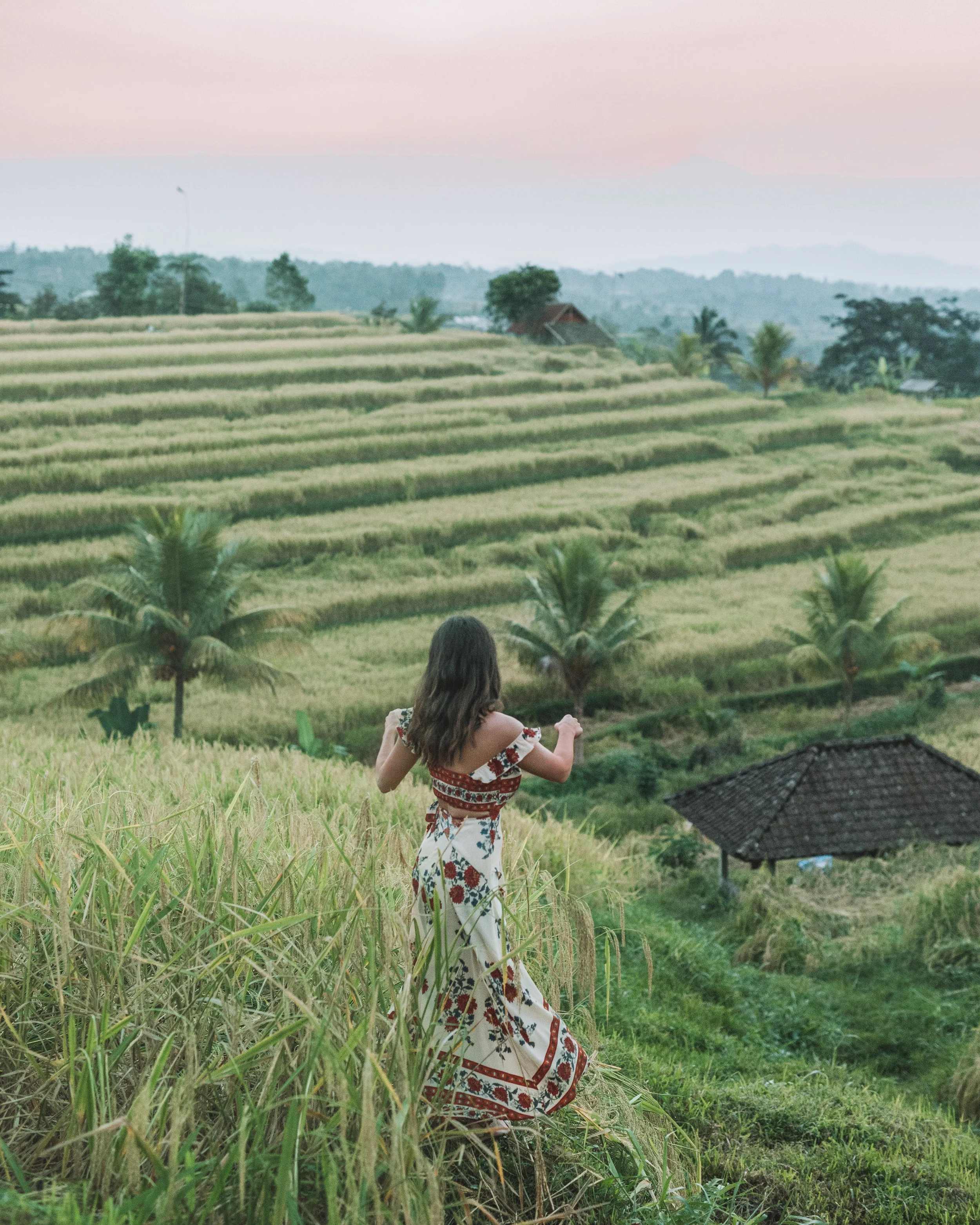 A woman in a floral dress standing in front of terraced rice fields at sunset with palm trees and a hut in the background.