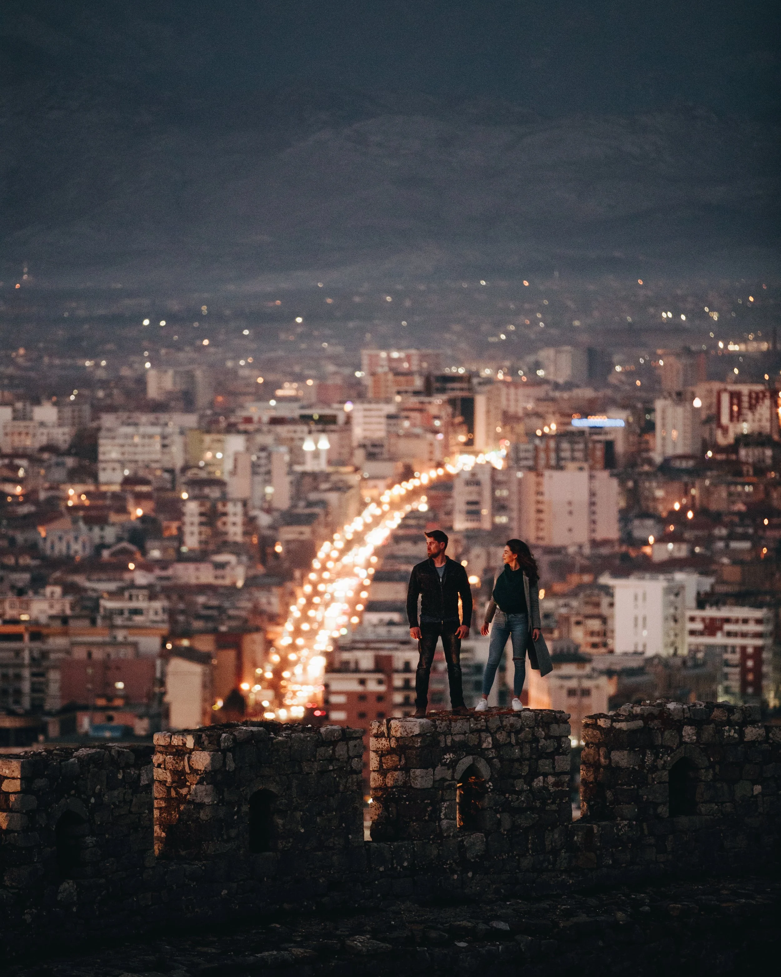 Two people standing on ancient stone ruins overlooking a city at night, with buildings and illuminated streets in the background.