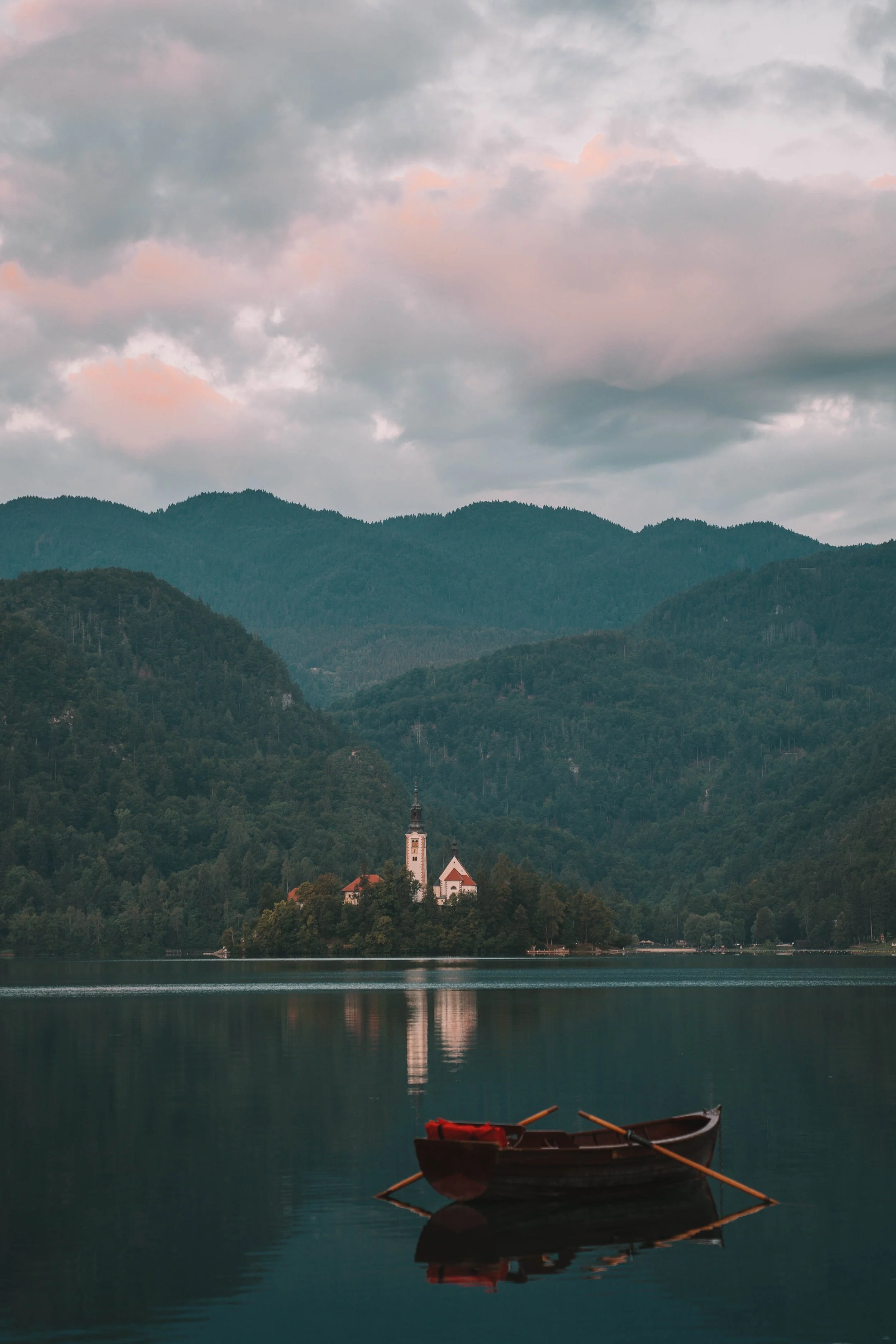 A serene lake with a small island featuring a church, surrounded by mountains and trees. A wooden rowboat with oars floats in the foreground under a cloudy sky.
