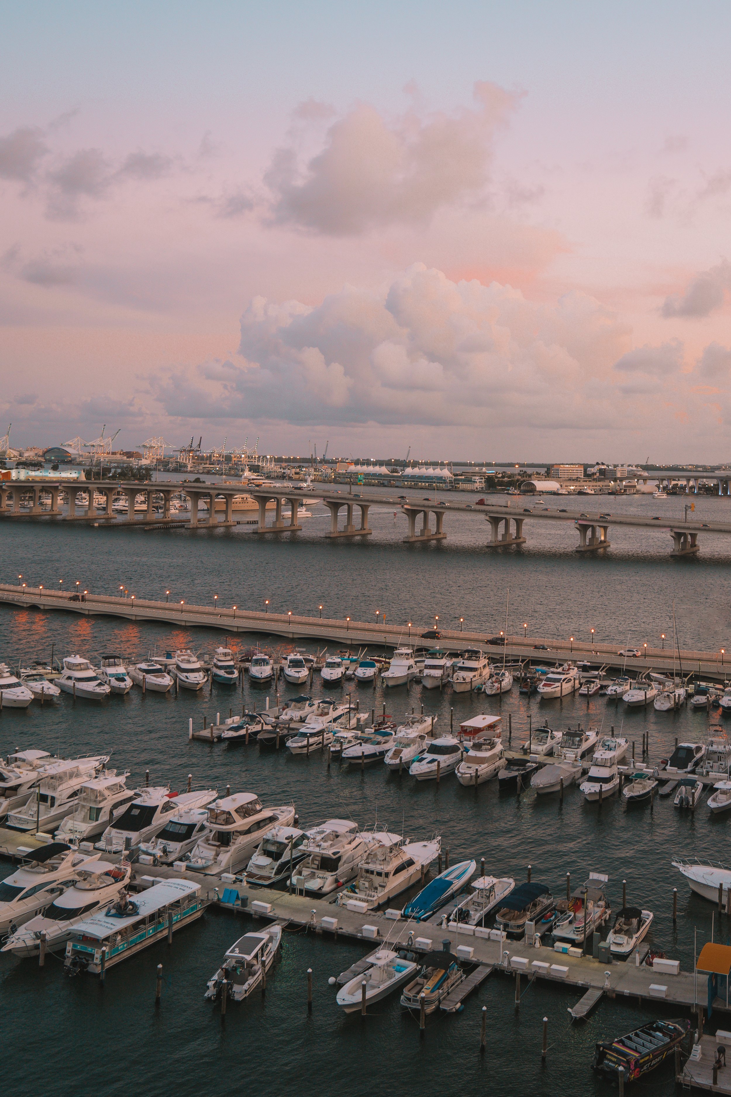 Marina with numerous boats docked, a bridge and cityscape in the background, under a cloudy sky at sunset.