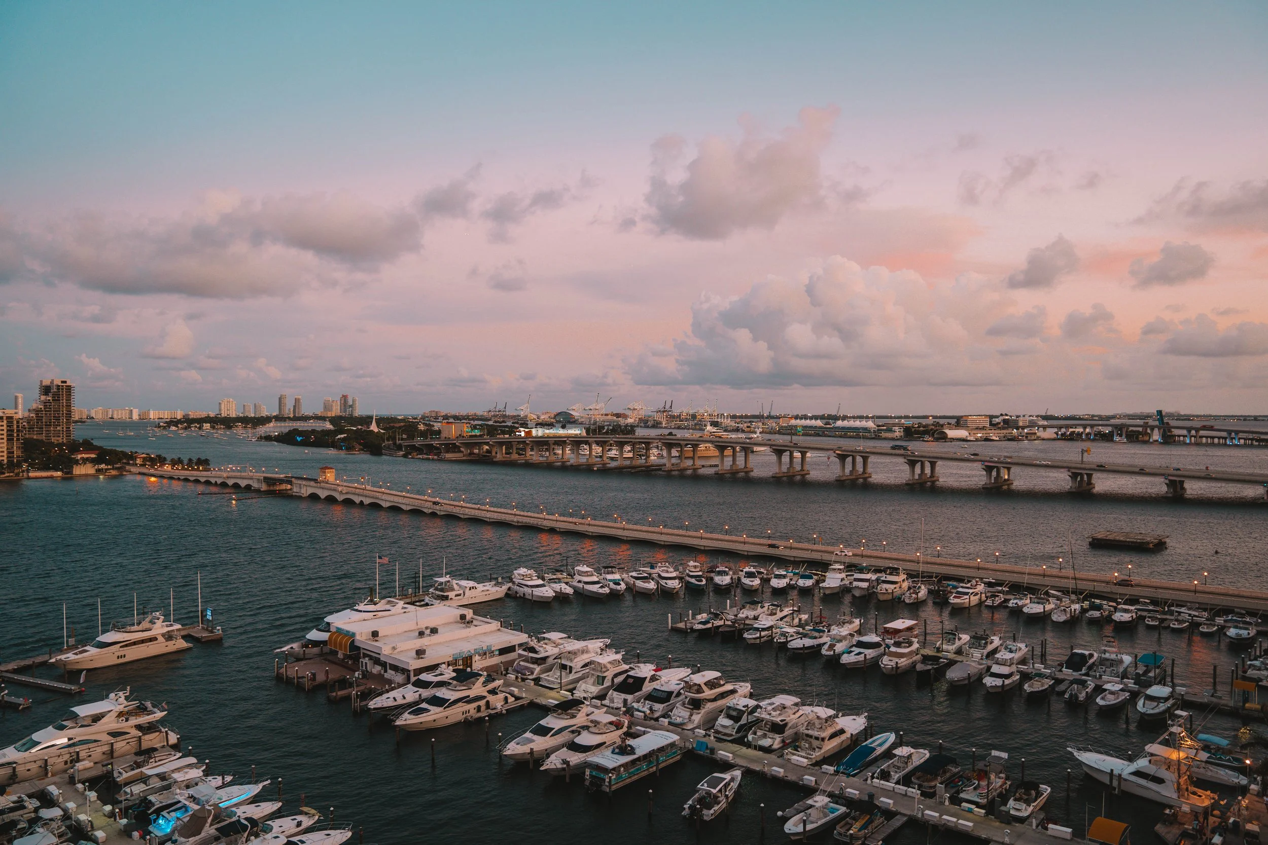 Marina with boats docked, bridge over water, city skyline, cloudy sky at sunset.