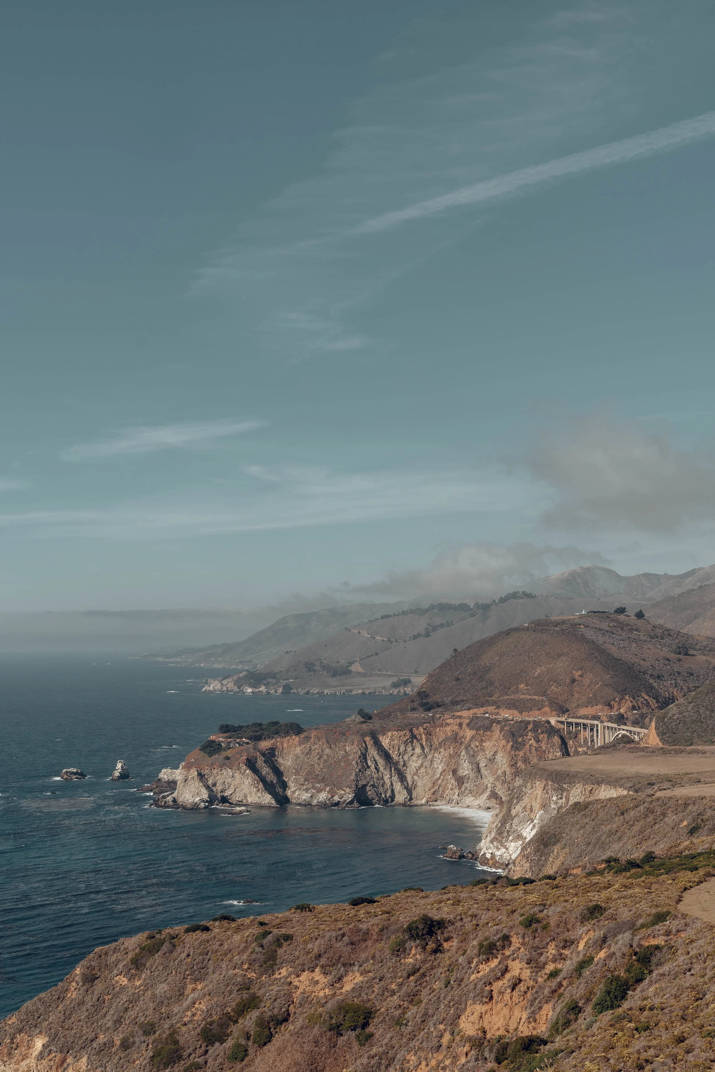 Scenic coastal view of cliffs, ocean, and a bridge under a blue sky.