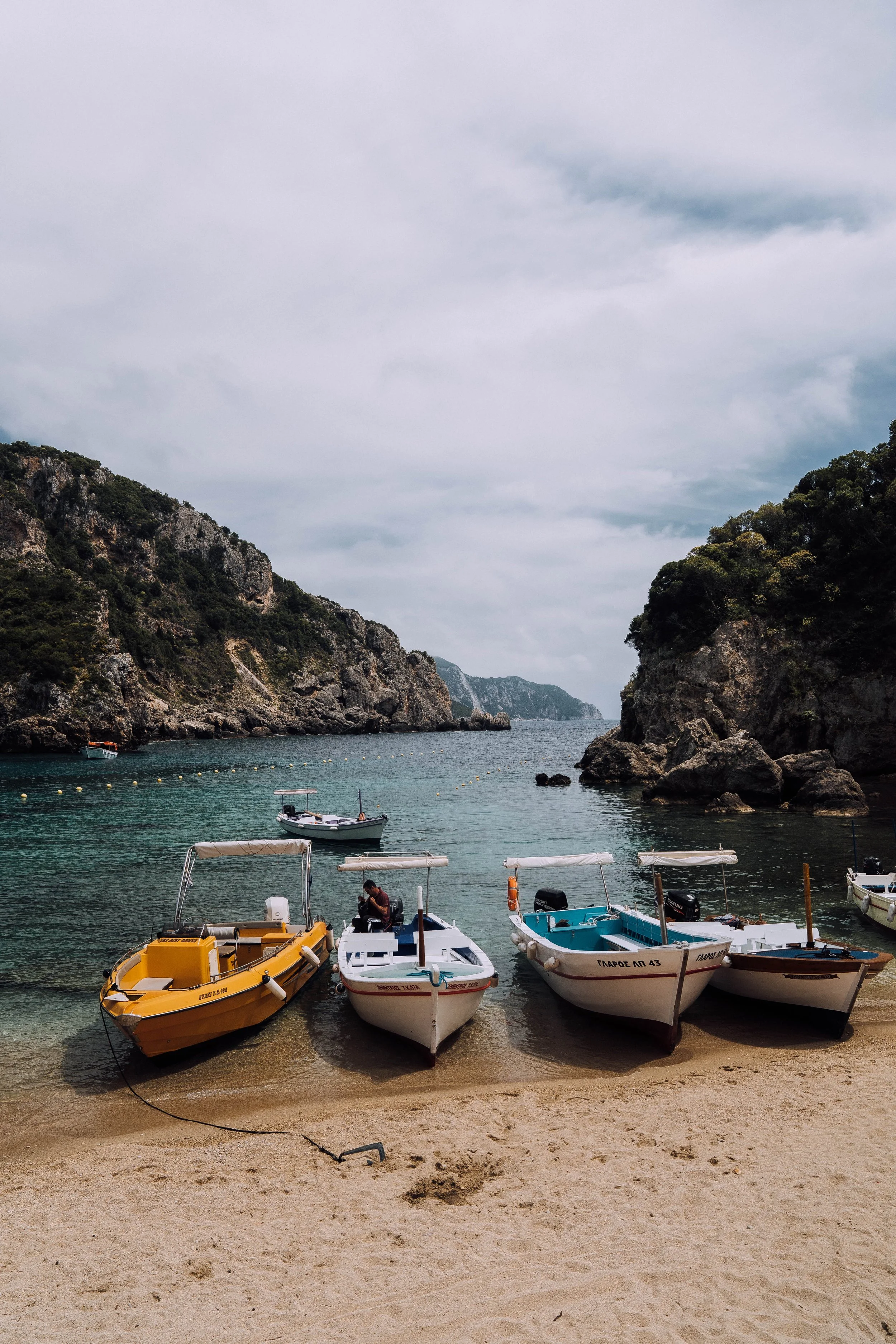 Four small boats lined up on a sandy beach with rocky cliffs and calm sea in the background.