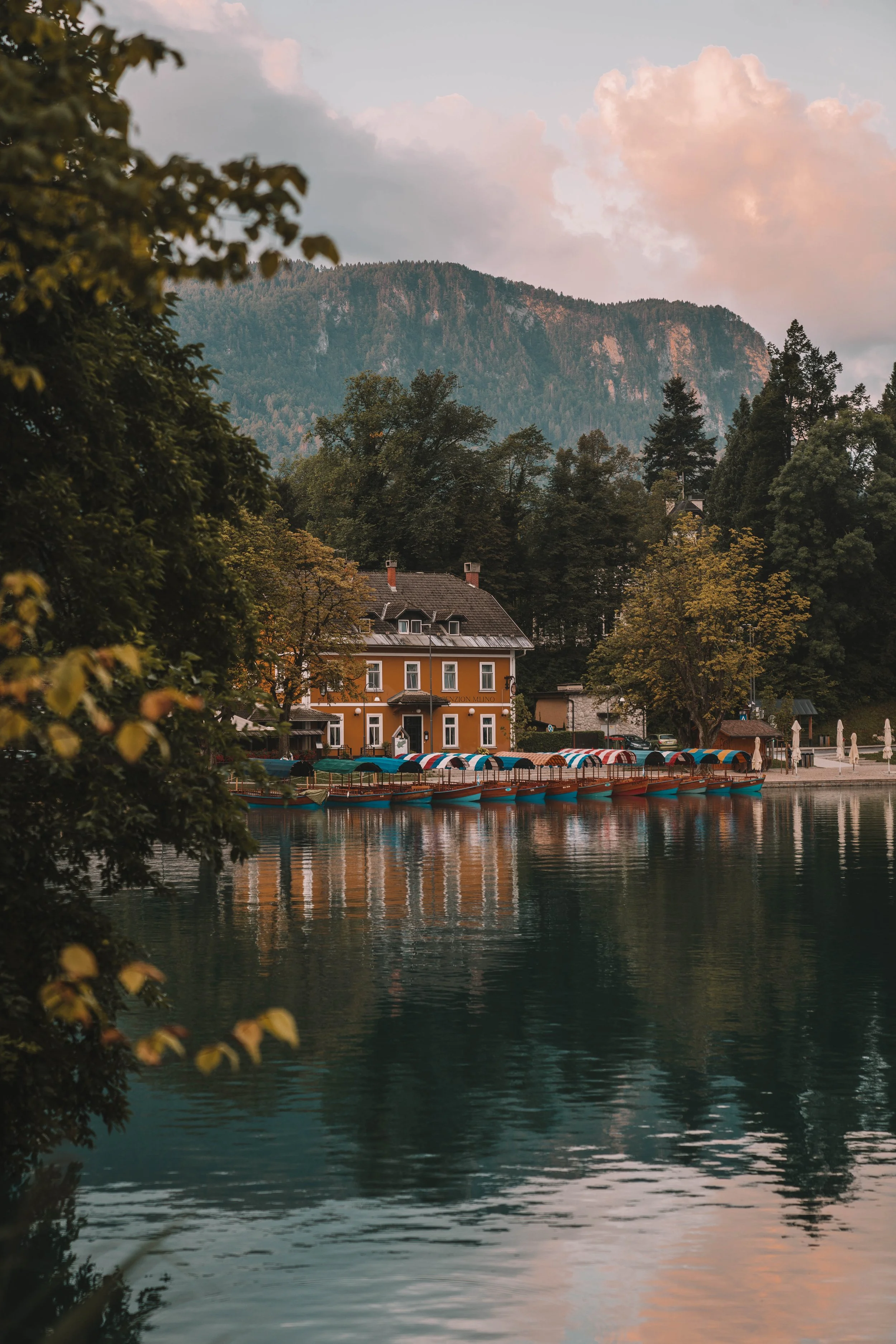 A lakeside house surrounded by trees, with colorful boats docked on the water and mountains in the background under a cloudy sky.
