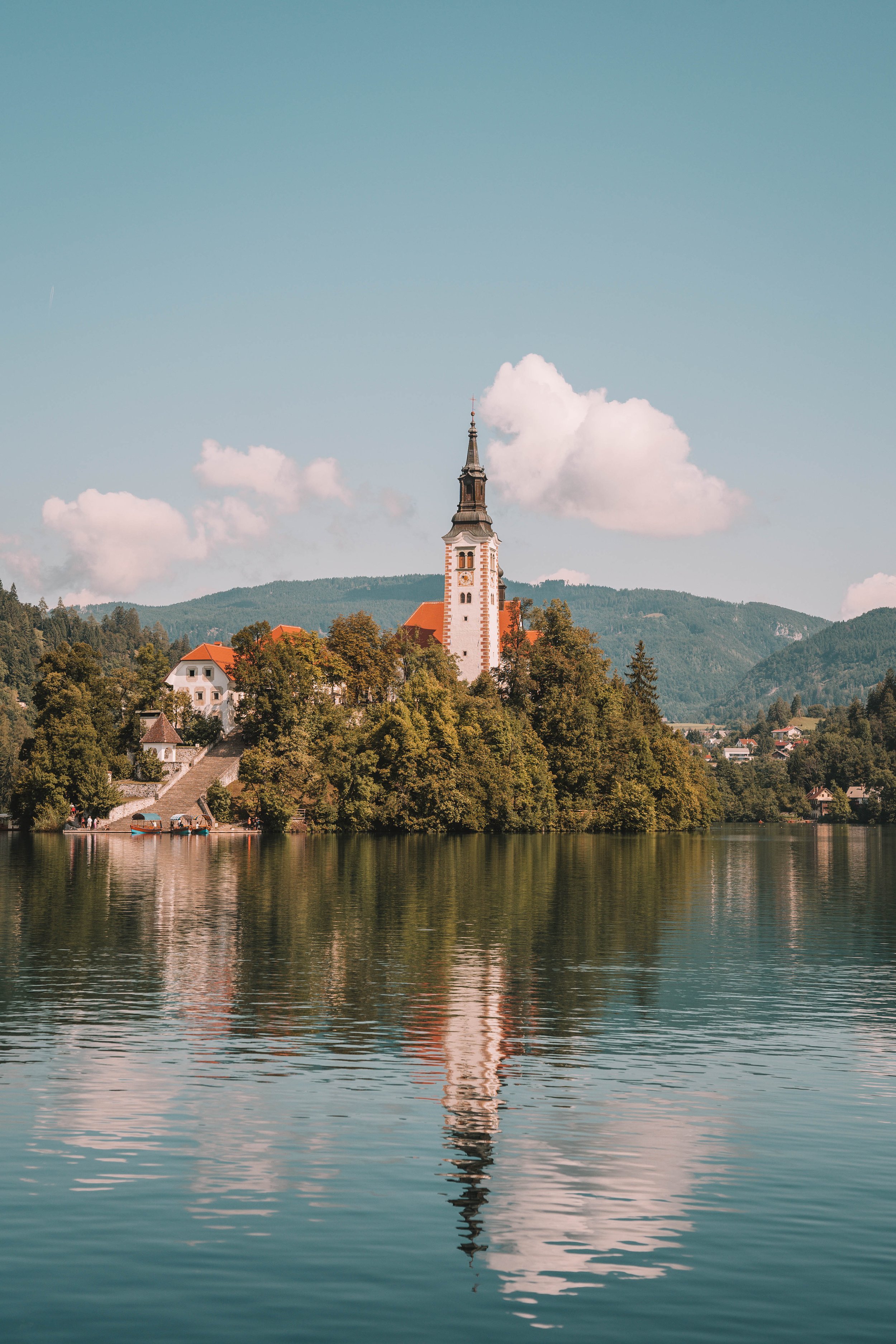 A picturesque church with a tall steeple surrounded by trees, located on an island in the middle of a calm lake, with reflections in the water and mountains in the background.