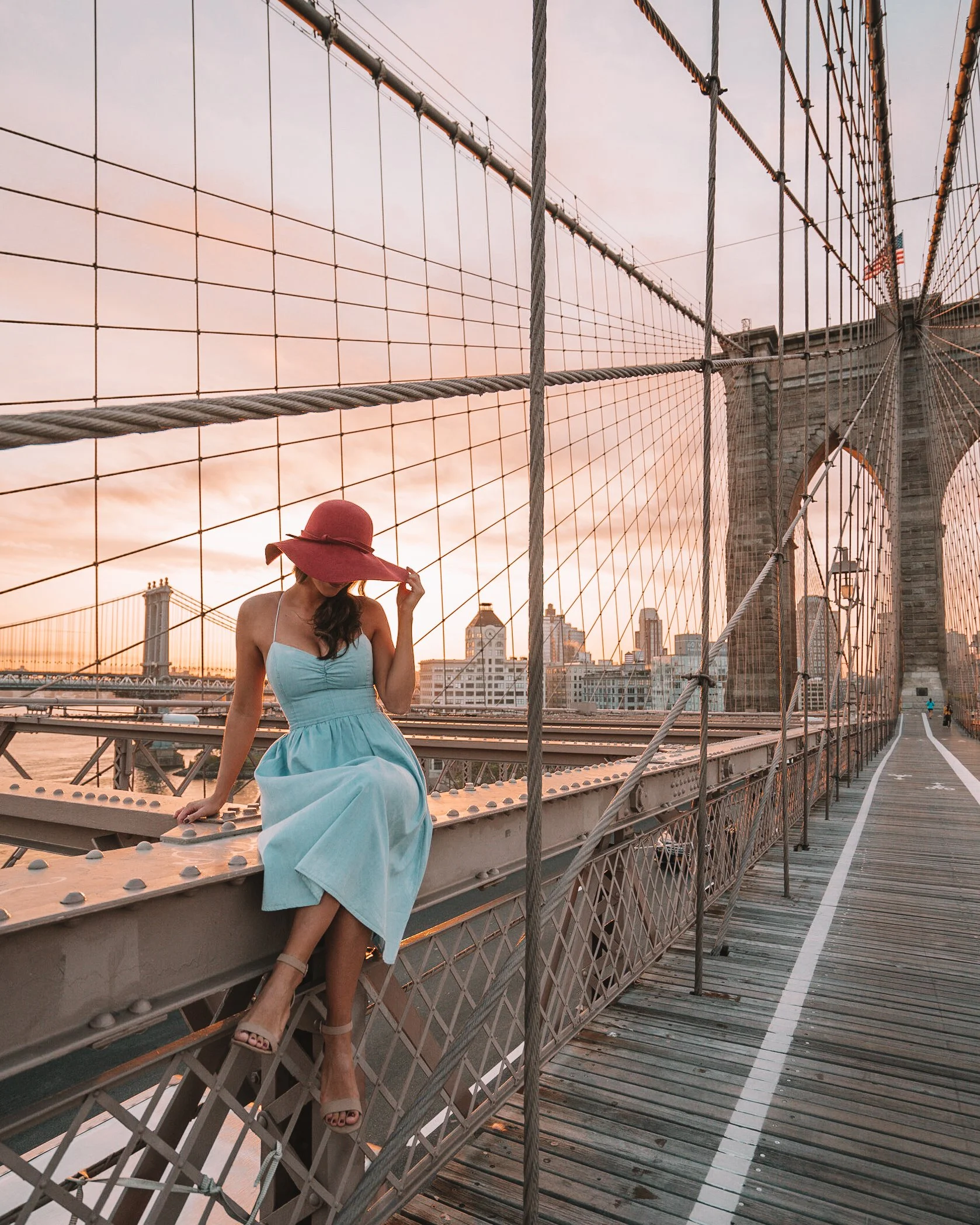Woman in blue dress and red hat sitting on railing of Brooklyn Bridge during sunset.