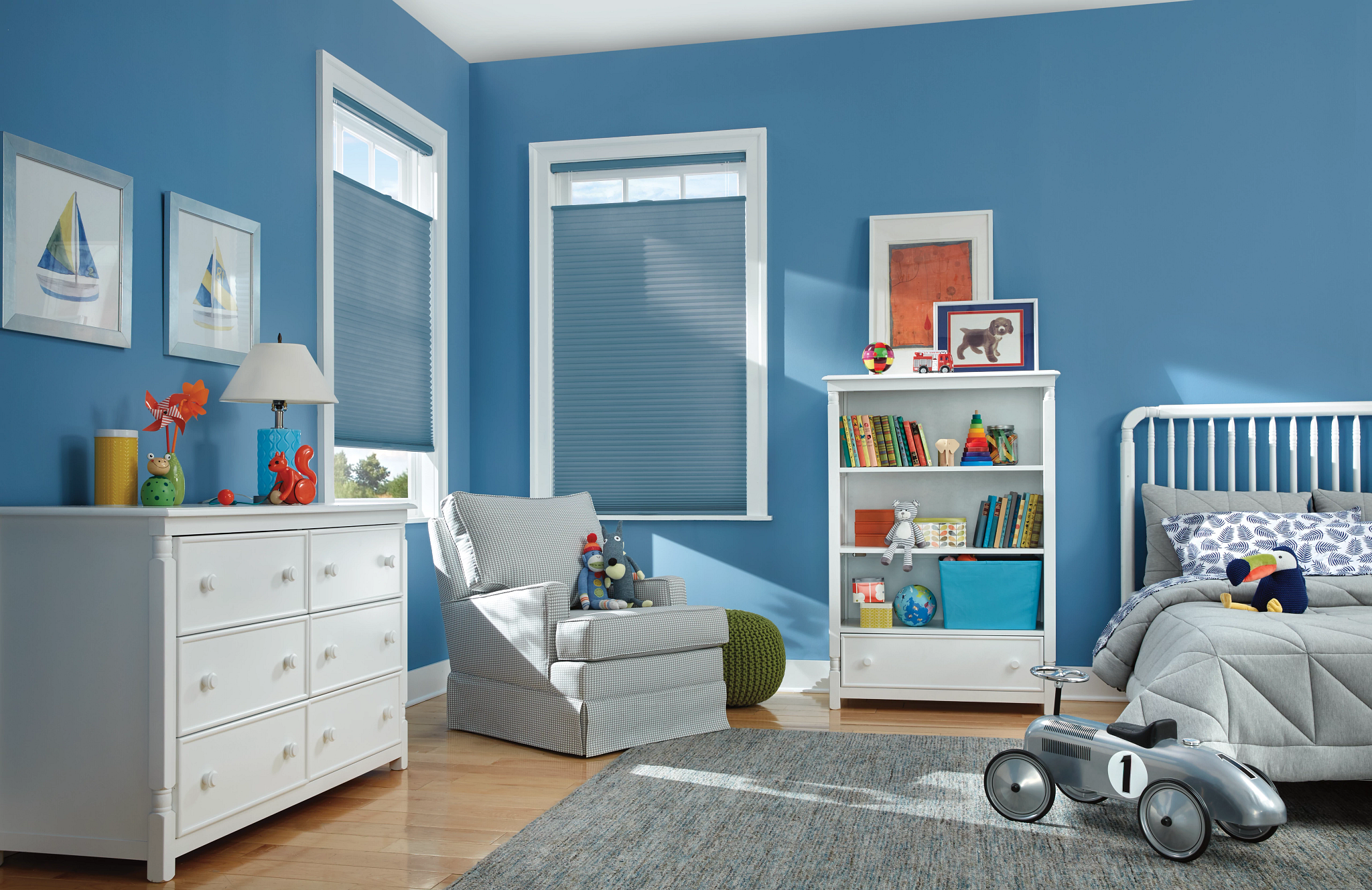 Children's bedroom with blue walls, white furniture, and toys, including a bed, bookshelf, armchair, and ride-on toy car.