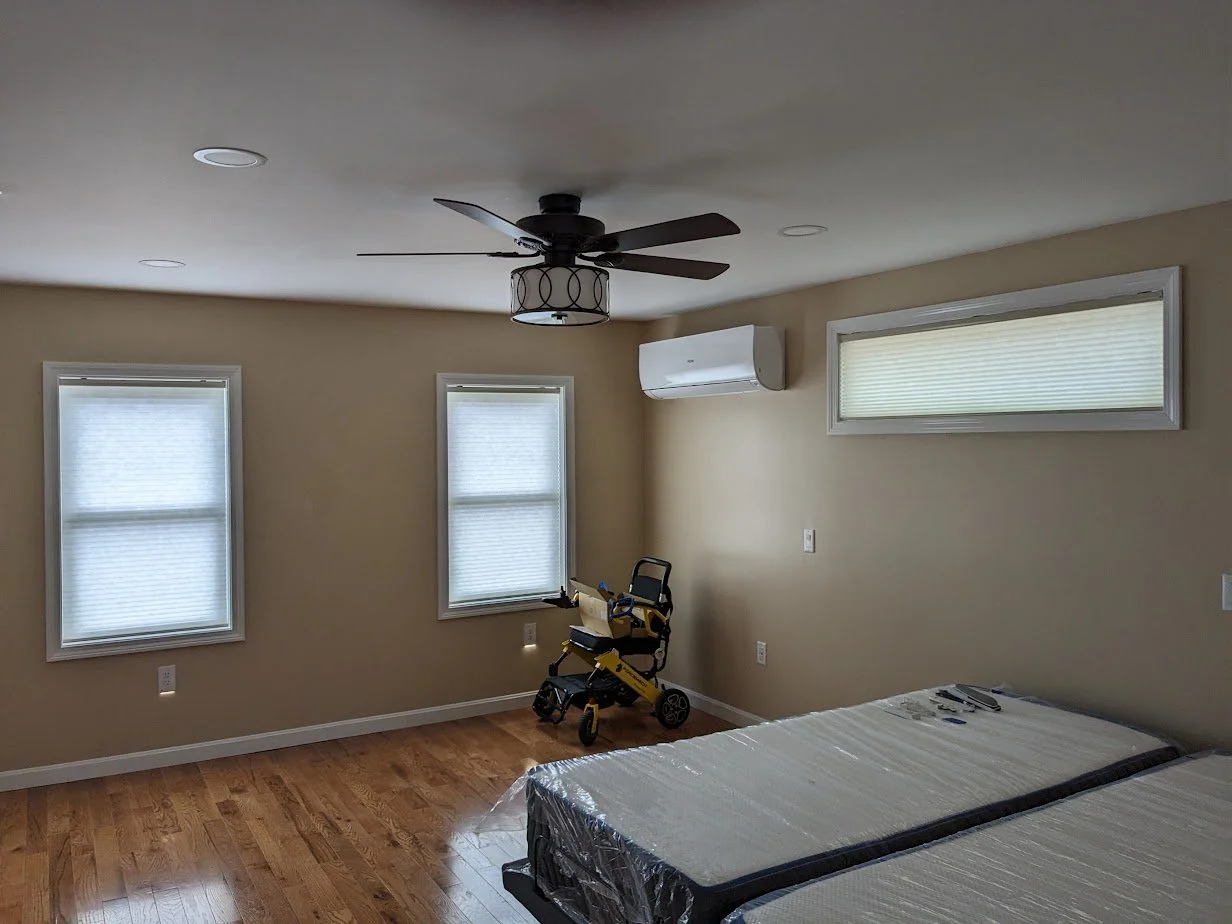 Empty bedroom with hardwood floors, three windows with blinds, a ceiling fan, wall-mounted air conditioning unit, and a mattress still in plastic packaging. There is a child's stroller next to the corner of the room.