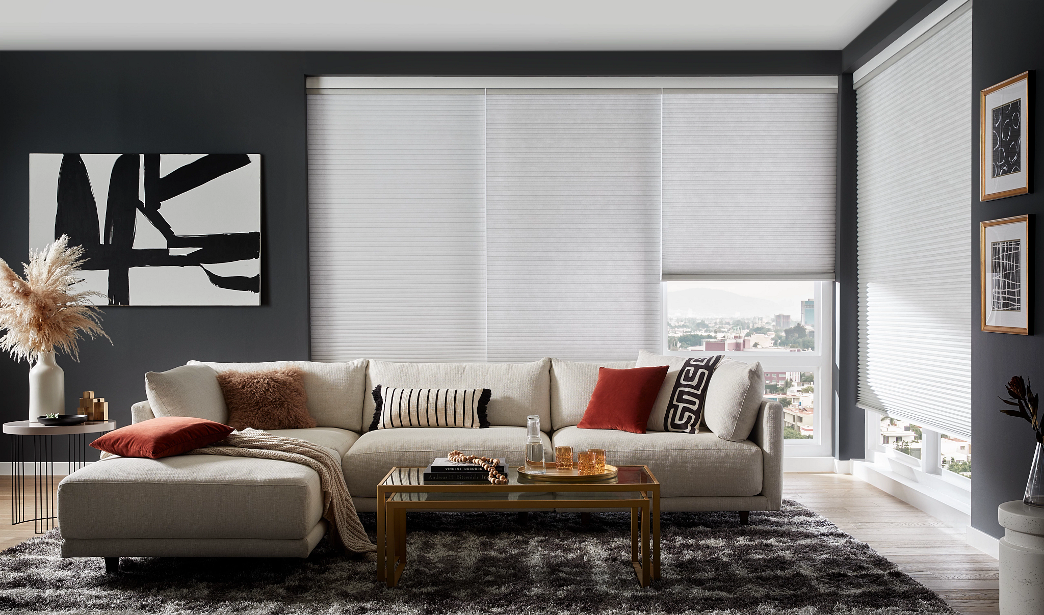 Modern living room with a white sectional sofa, red and patterned pillows, black and white wall art, window blinds, and city view