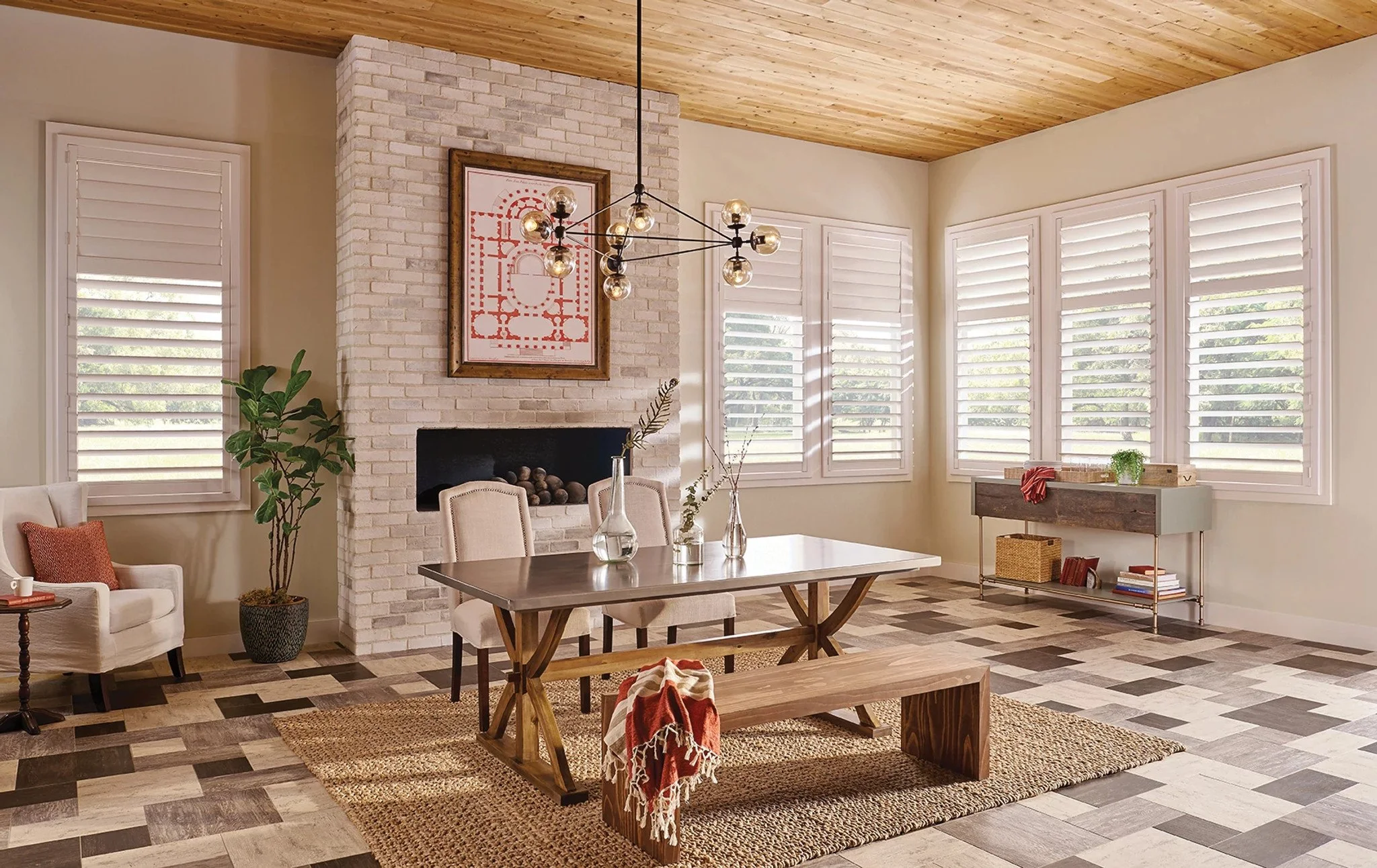 Living room with a brick fireplace, wooden ceiling, multiple windows with shutters, and modern decor including a white armchair, potted plant, wooden table, glass vases, and a bench with a throw blanket.