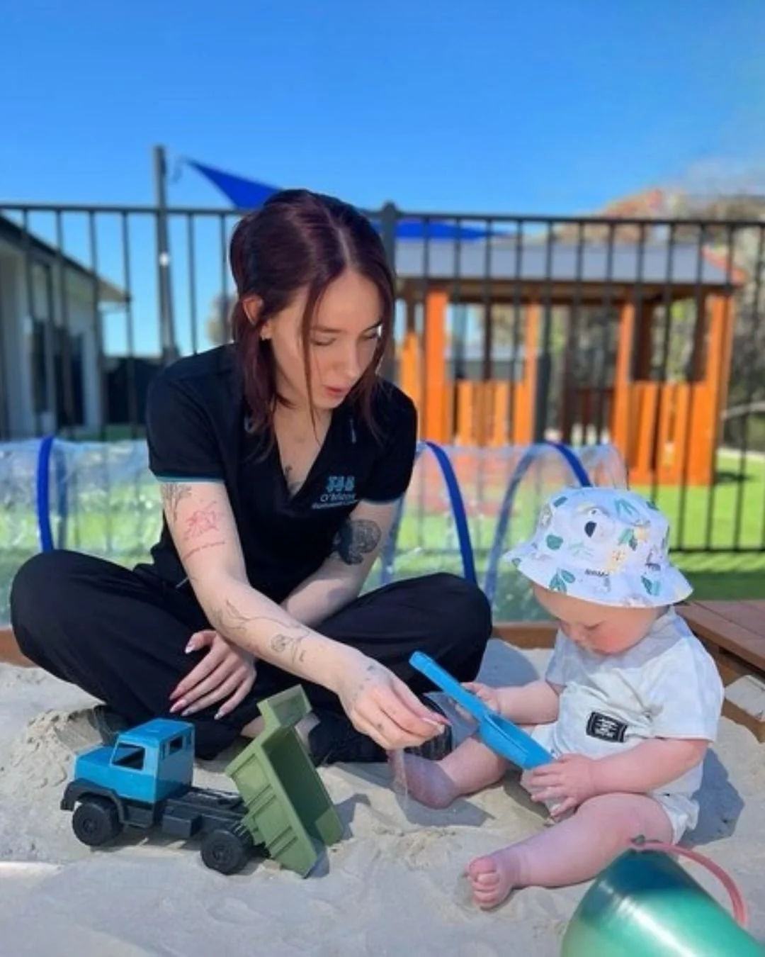 A woman with tattoos on her arms sitting on sand and playing with a young child wearing a white hat with animal illustrations, in a backyard with toys, a wooden playset, and a metal fence under a clear blue sky.