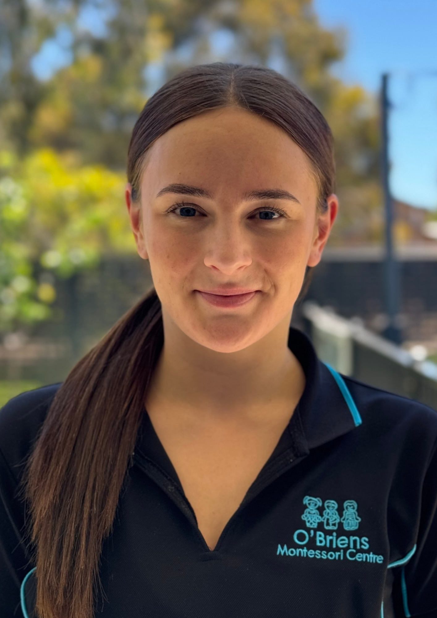 A young woman with long brown hair smiling outdoors, wearing a black uniform with the logo and text of O'Brien's Montessori Centre.