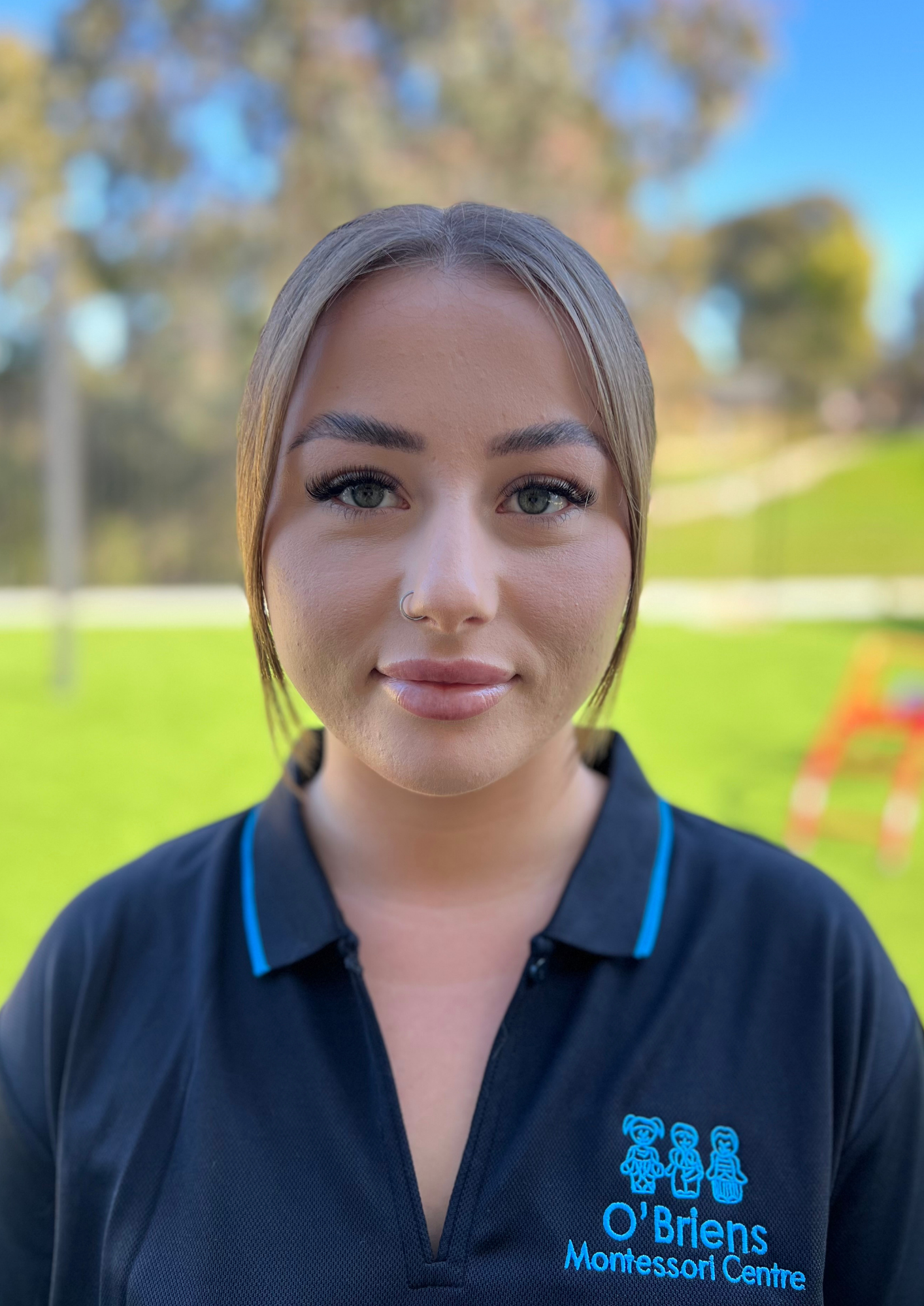 Close-up of a young woman with blonde hair and gray eyes, wearing a black collared shirt with blue accents and the logo of O'Brien Montessori Centre in blue, outdoors with a background of trees and a green field.