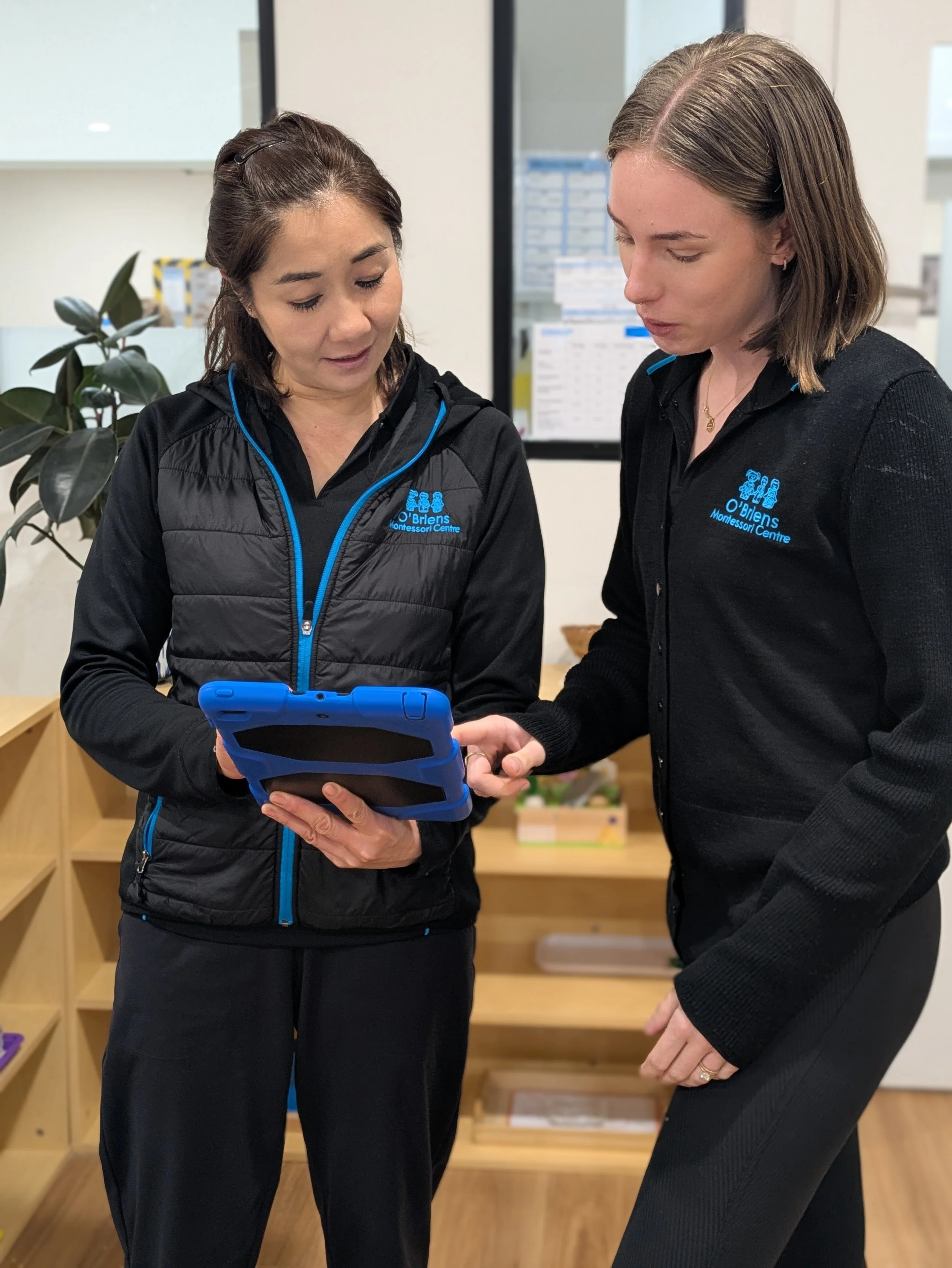 Two women, one with dark hair and the other with blonde hair, looking at a handheld device together in an indoor setting with light-colored walls and wooden shelves.
