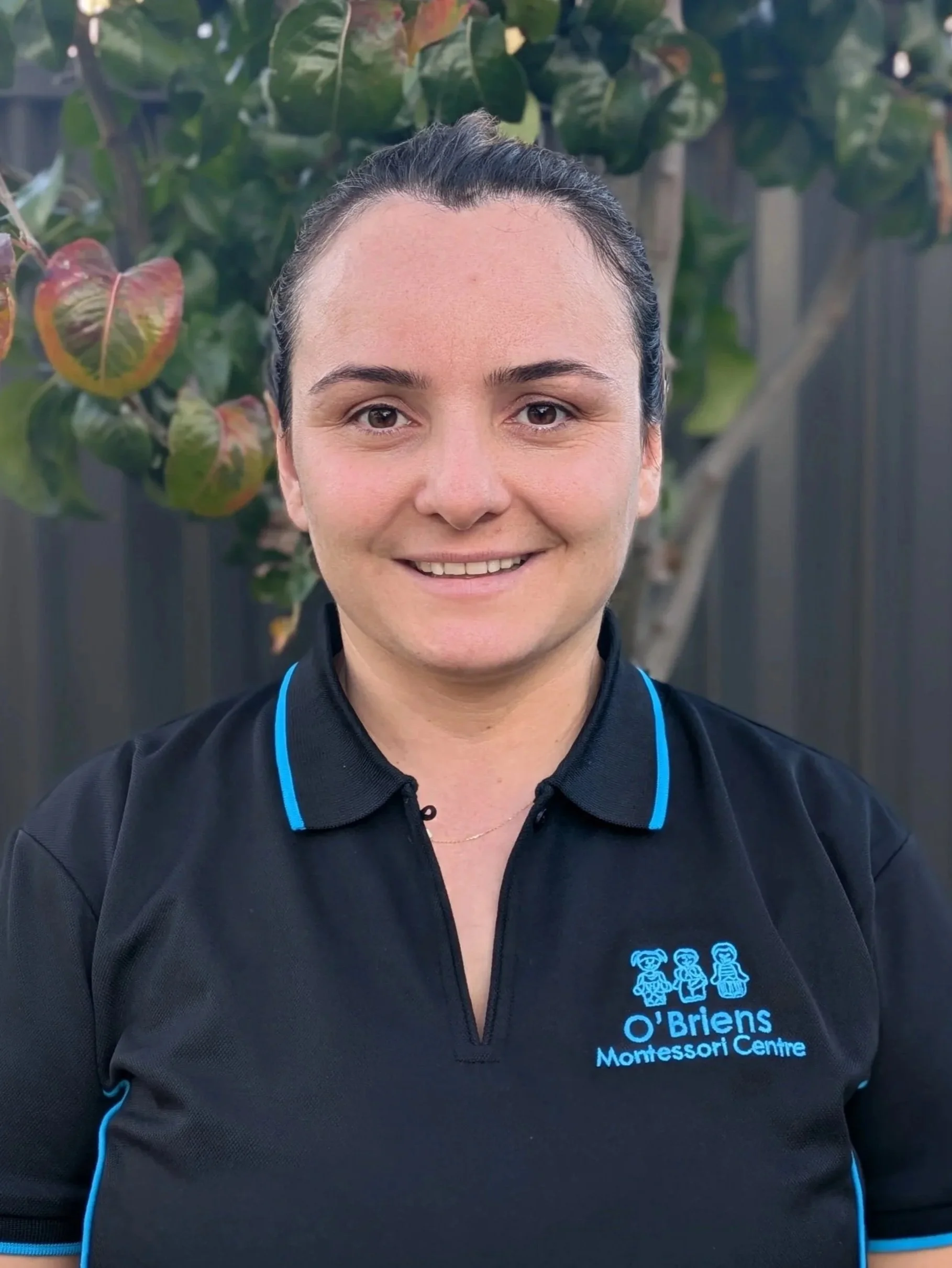 Close-up of a woman in a black polo shirt with blue accents, smiling outdoors with green leaves in the background. The shirt has the logo of 'O'Brien's Montessori Centre' featuring three cartoon children.