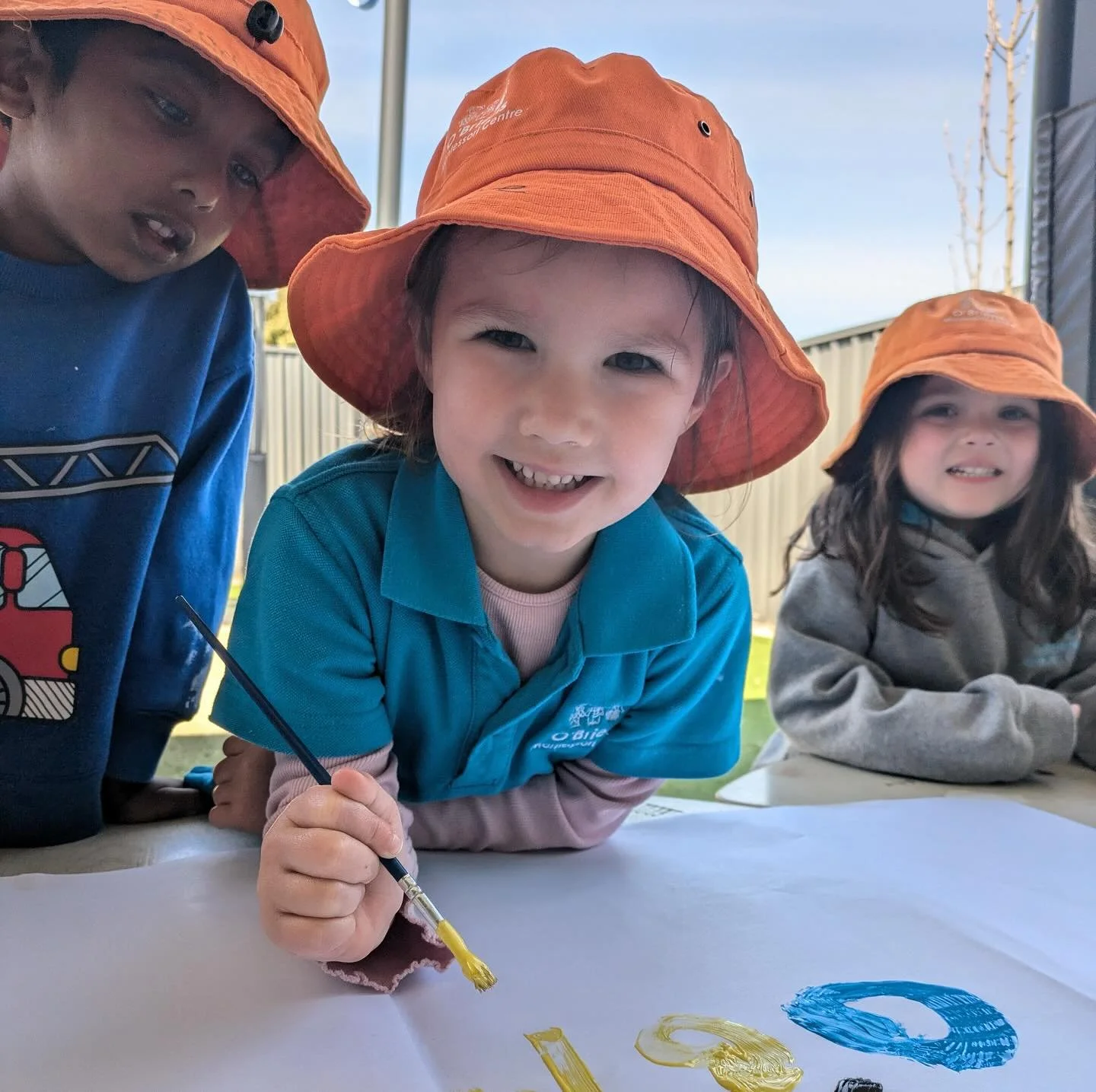 Three children wearing orange hats and colorful shirts are leaning over a table, smiling at the camera while painting on a large white sheet of paper outdoors.