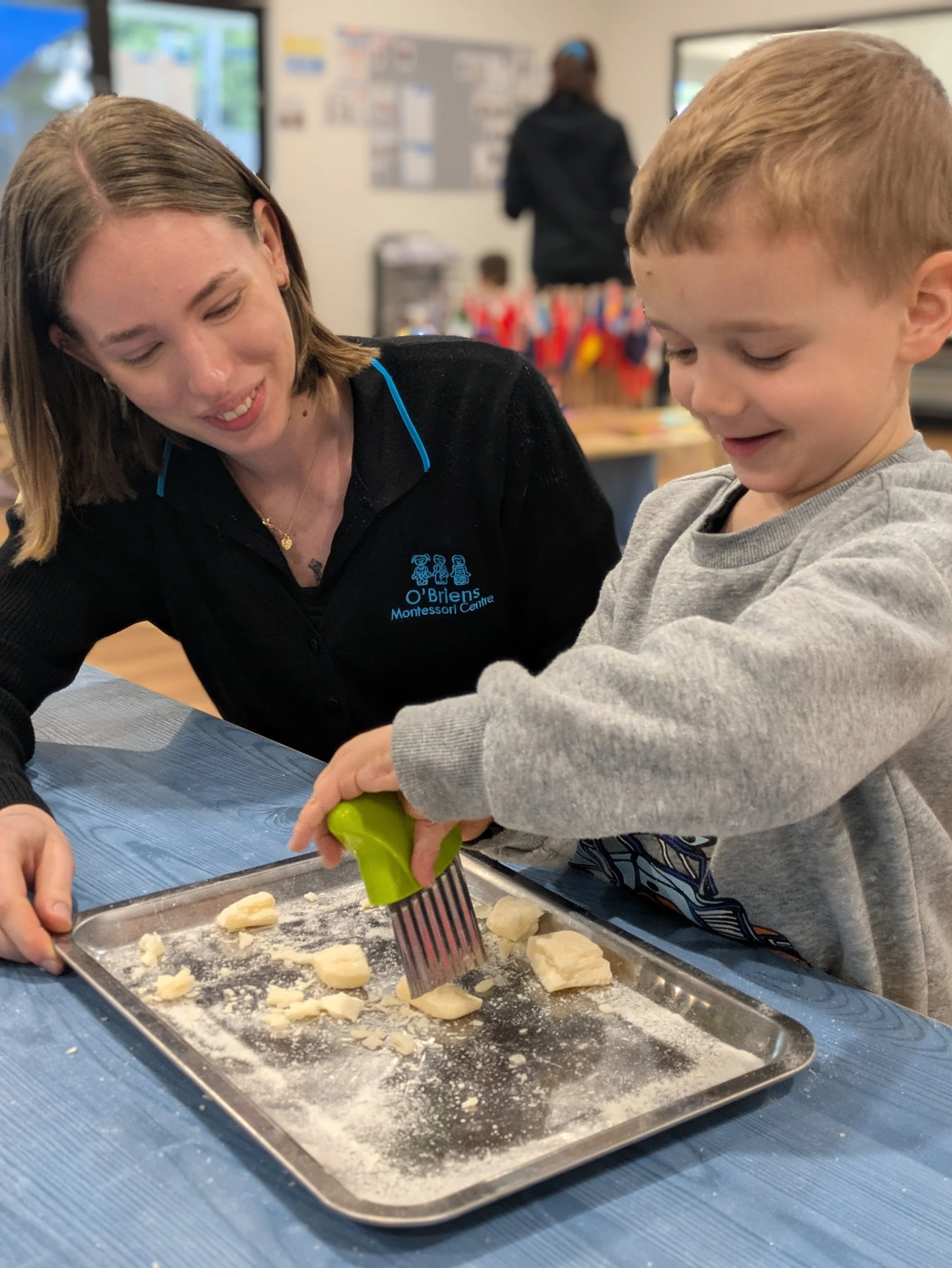 A woman and a young boy preparing dough on a baking sheet in a classroom setting.