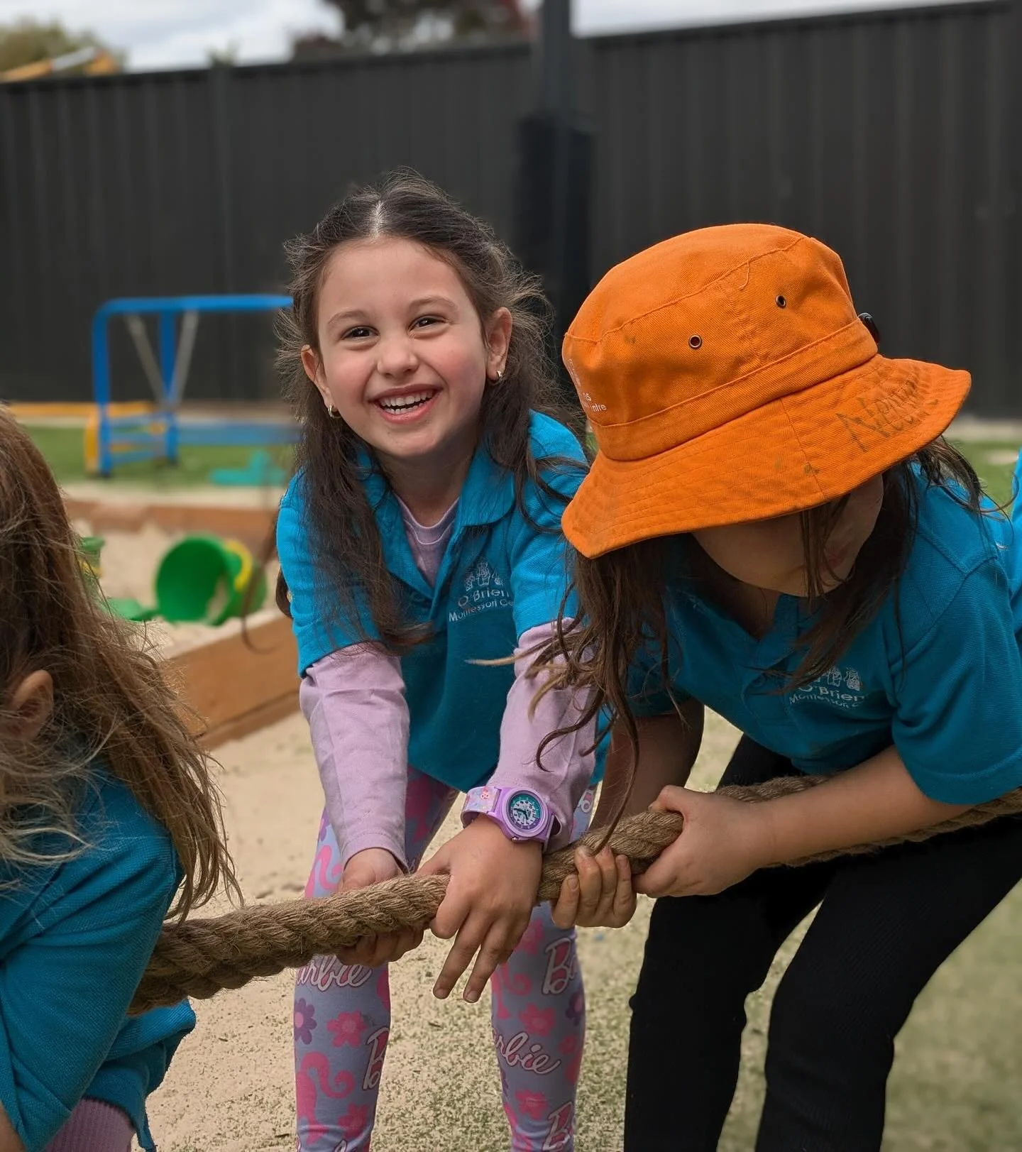 Young girl with a big smile playing tug-of-war with others in a playground sandpit, wearing a pink watch, blue shirts, and a large orange hat, with playground equipment in the background.