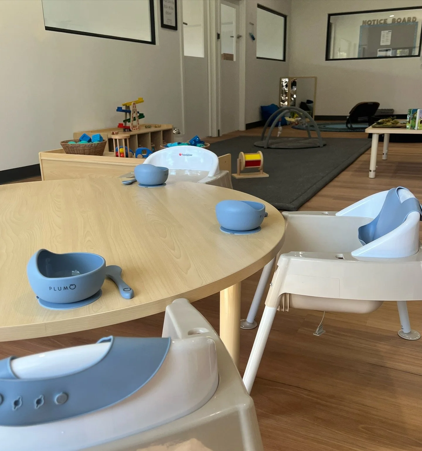 Nursery or daycare room with a round wooden table with blue placemats and bowls, white high chairs, and various toys and play mats in the background.