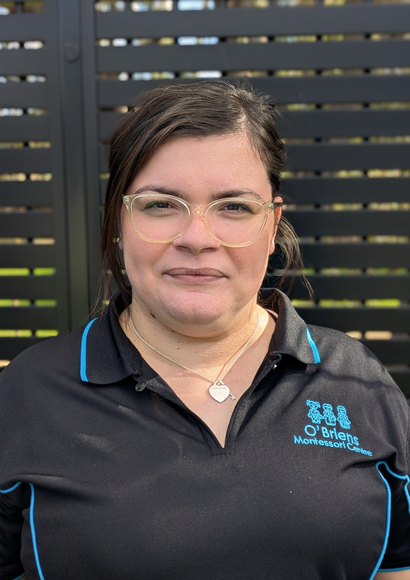 A woman with dark hair and glasses wearing a black polo shirt with blue accents and the O'Brien Montessori Centre logo, standing outdoors in front of a black slatted fence.