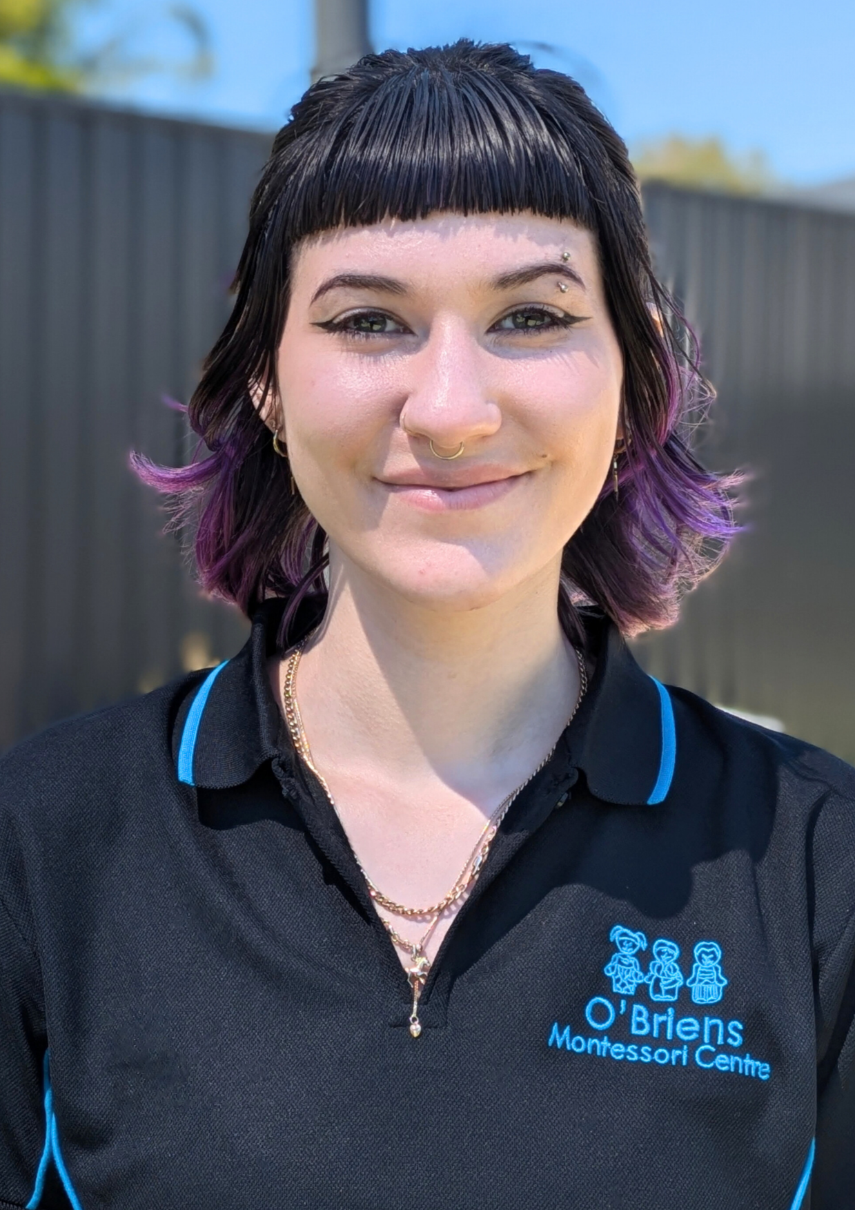 A woman with short black hair and purple tips, wearing a black shirt with blue accents and logo that reads 'O'Brien Montessori Centre'. She has piercings, including a nose ring and eyebrow rings, and is wearing necklaces. She is outdoors with a blurred background.