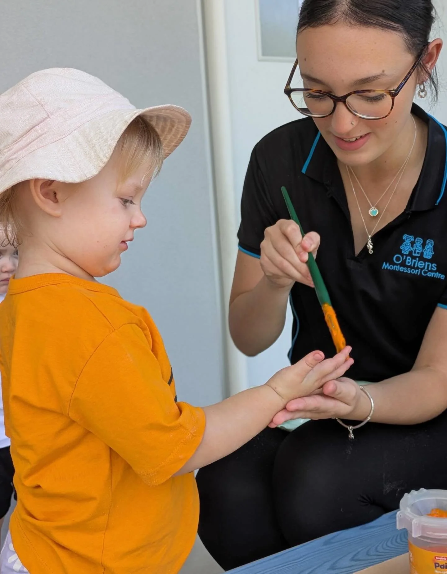 A young boy wearing an orange shirt and a sun hat receives a manicure from a smiling woman wearing glasses and a black polo shirt with a logo. The woman is holding the boy's hand and applying polish or paint to his fingernails with a green brush.