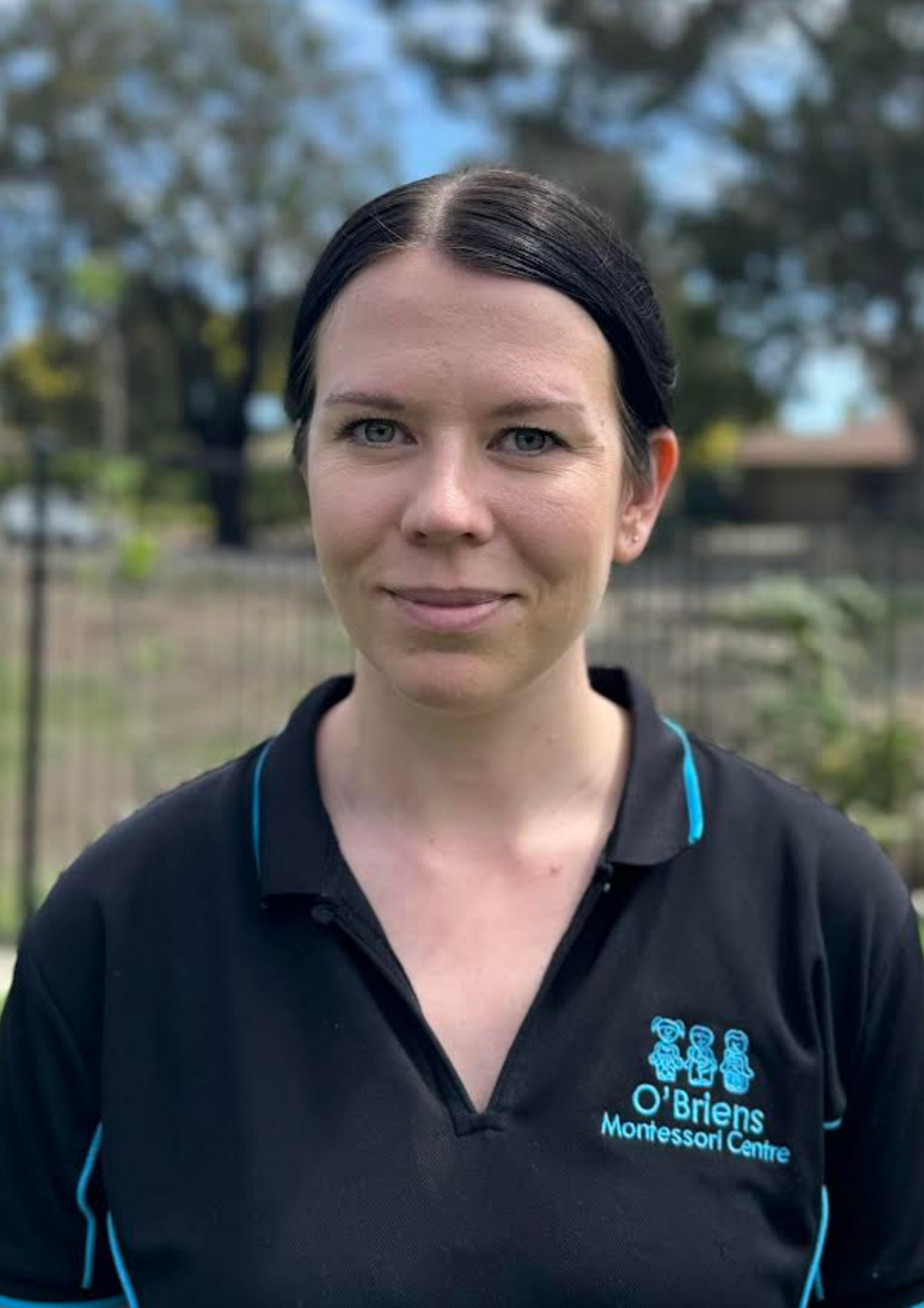 Close-up of a woman with dark hair in a ponytail, wearing a black polo shirt with the O'Brien Montessori Centre logo, outdoors with trees and a fence in the background.