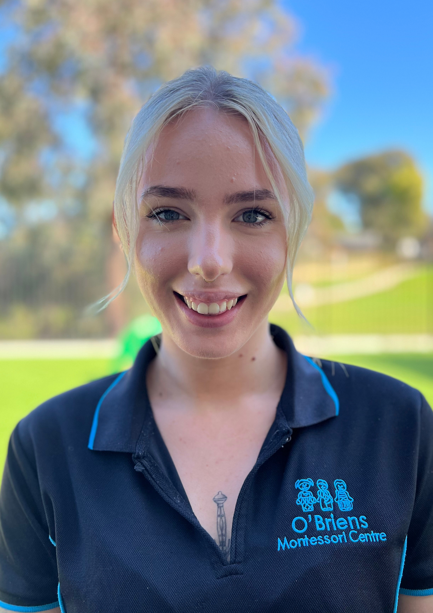 Close-up of a smiling young woman with blue eyes, blonde hair, and freckles, wearing a black polo shirt with blue accents and logo of O'Brien's Montessori Centre, outdoors on a sunny day with blurred trees in the background.