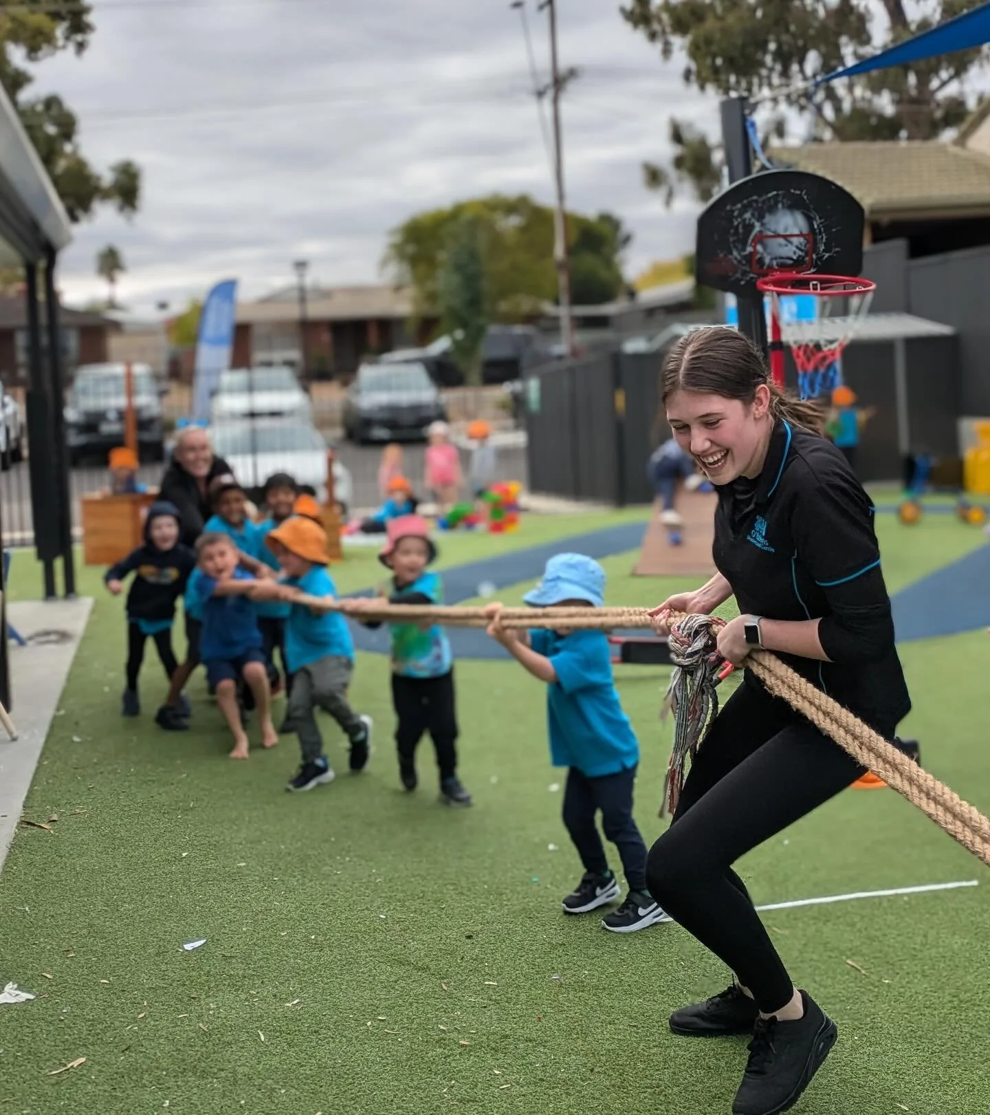 Group of children and an adult participating in a tug-of-war game on an outdoor playground during daytime, with a basketball hoop and a fence in the background.