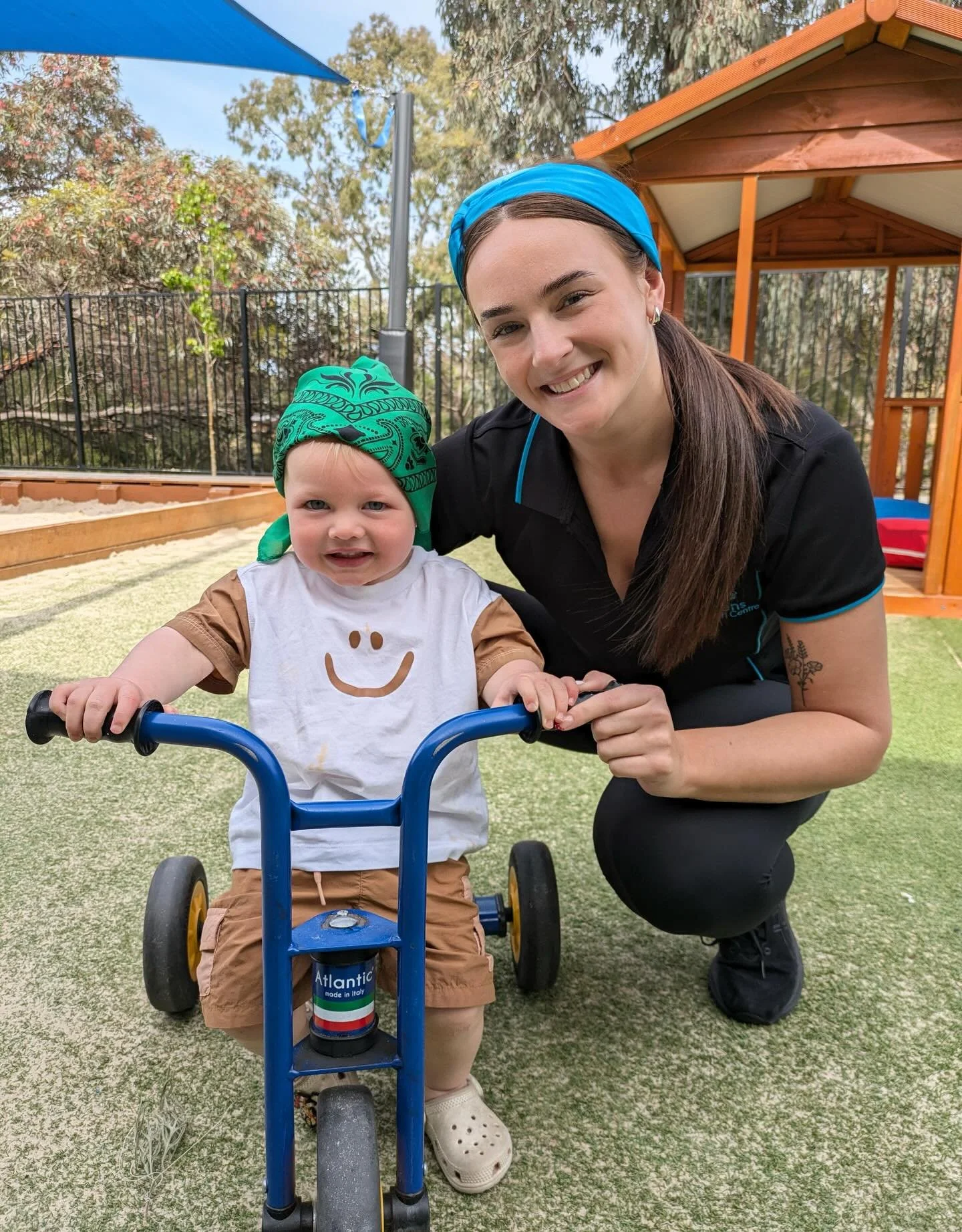 A young woman with long brown hair, wearing a blue headband and a black shirt, kneels next to a toddler on a tricycle, smiling outdoors in a backyard area with trees, a red-roofed wooden structure, and a fenced garden in the background.