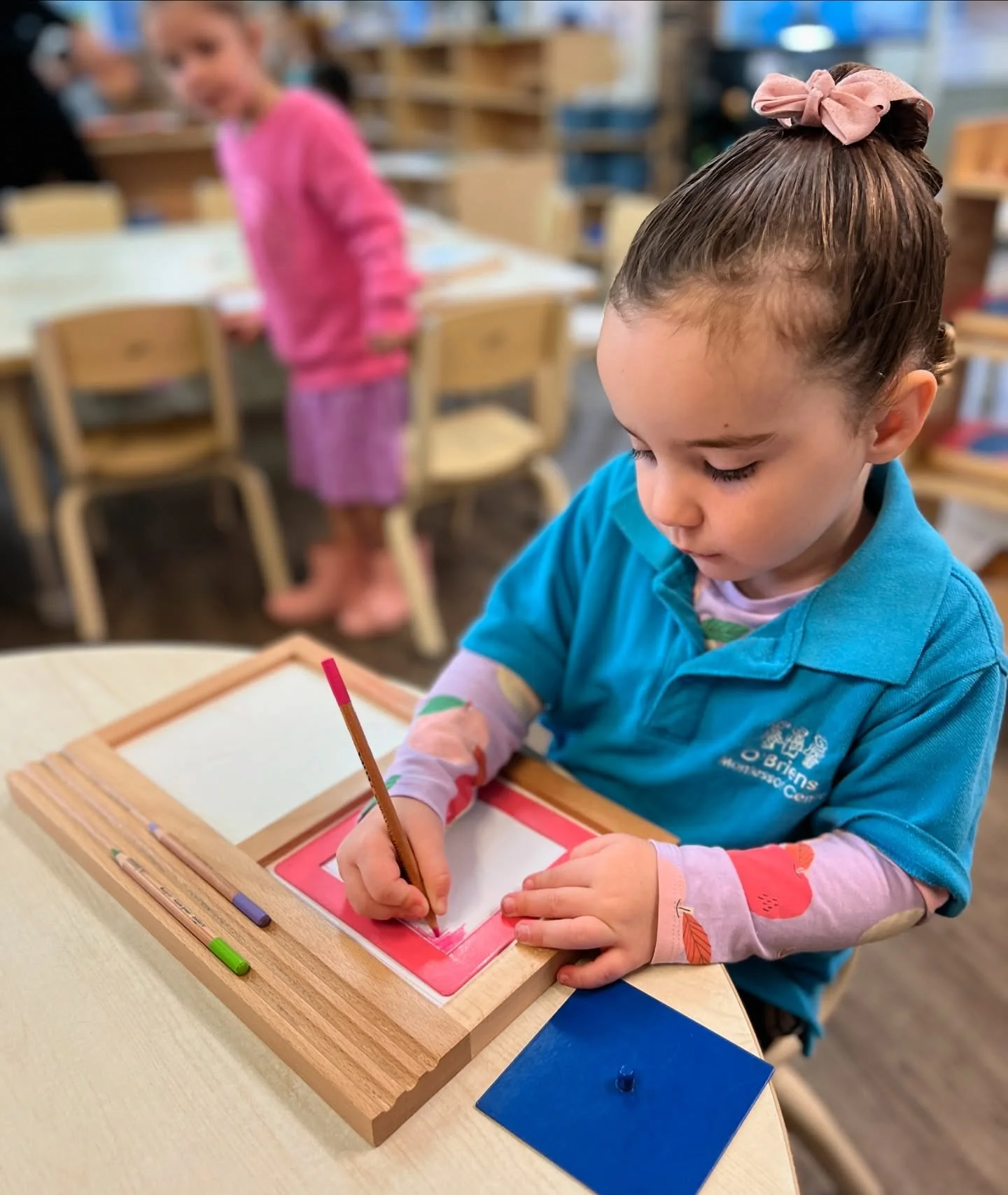 A young girl in a blue shirt and pink long sleeve shirt painting on a small canvas with a paintbrush at a table in a classroom. A girl in a pink sweater and skirt is blurred in the background.