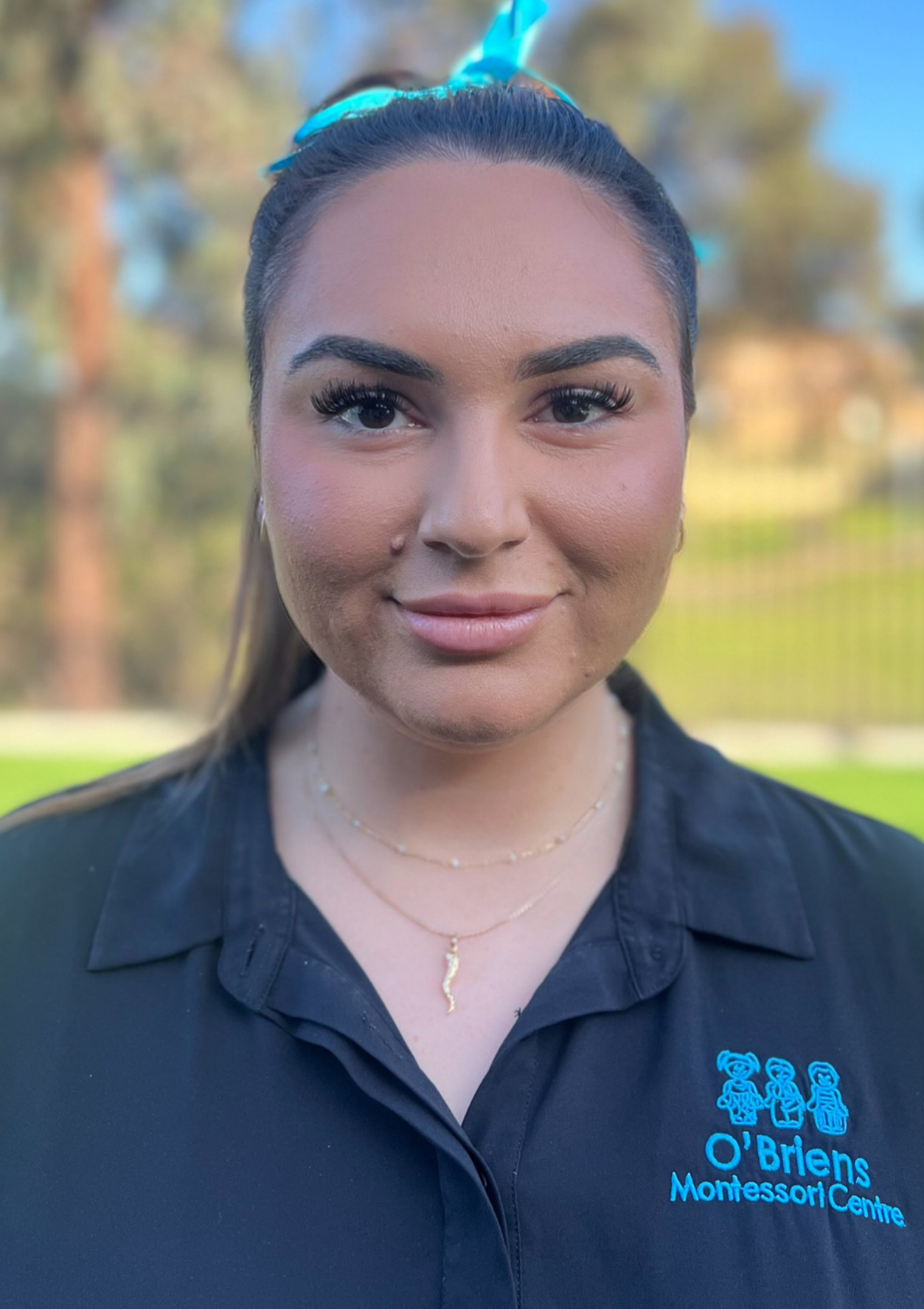 Close-up of a woman with dark hair, wearing a blue hair bow, black shirt, and layered necklaces, standing outdoors with blurred trees in the background. She has makeup and is smiling slightly. The shirt has a logo of children and text that reads "O'Brien Montessori Centre."