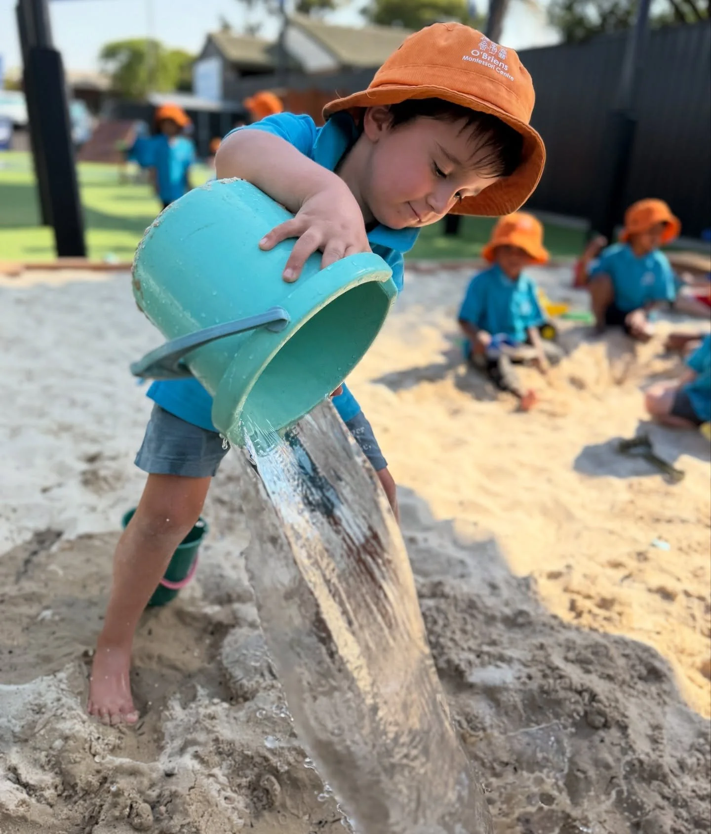 Young boy wearing an orange hat and blue shirt pouring water from a blue bucket onto sand in a playground or sandpit, with other children and a fence in the background.