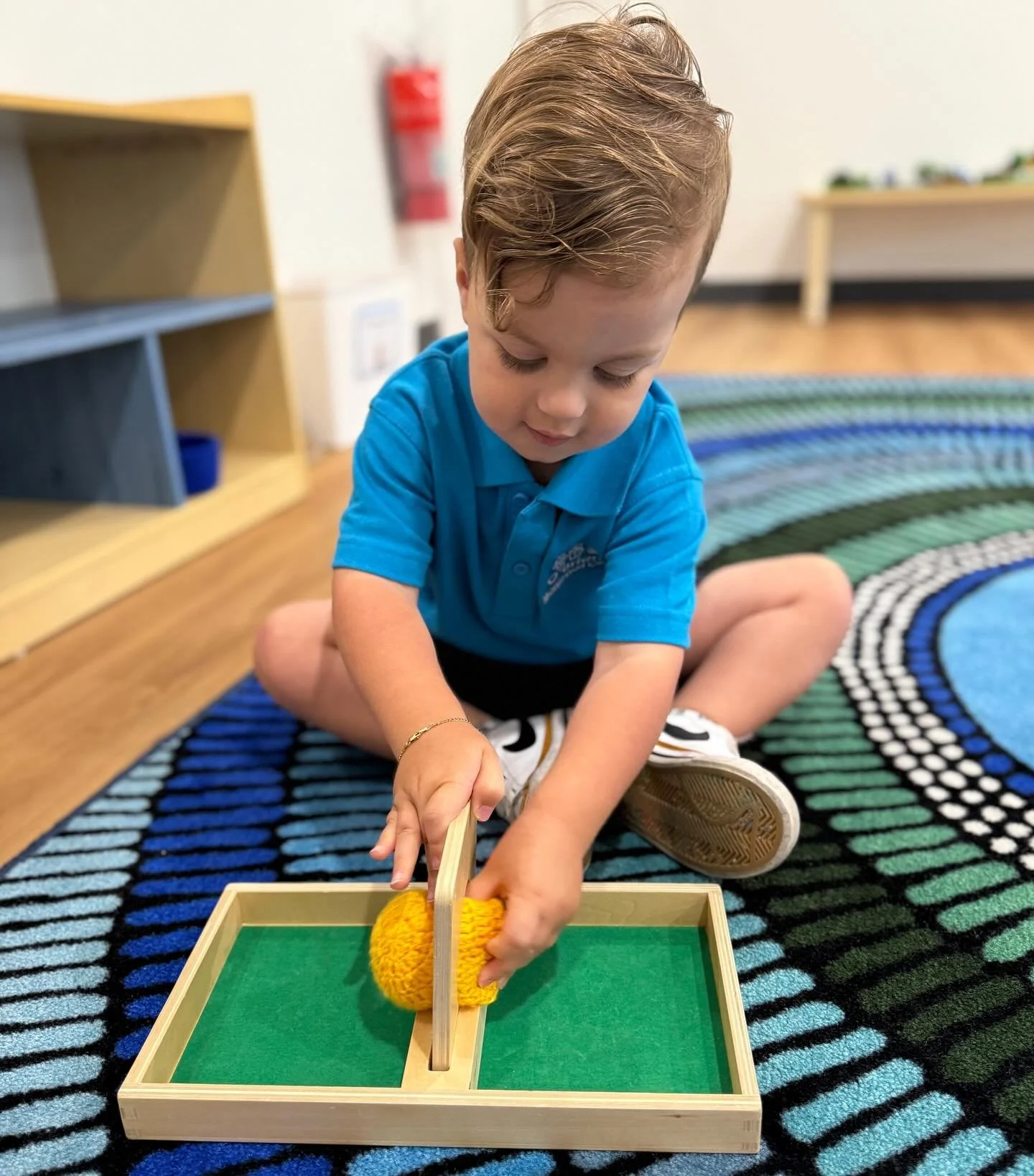 Young boy sitting on a colorful striped rug in a classroom, playing with a wooden toy digging game involving a yellow sponge and a small shovel.