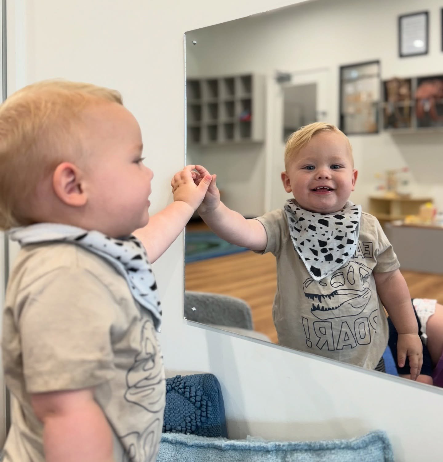 A young boy with blond hair smiling at his reflection in a mirror, touching his hand to his reflection's hand.