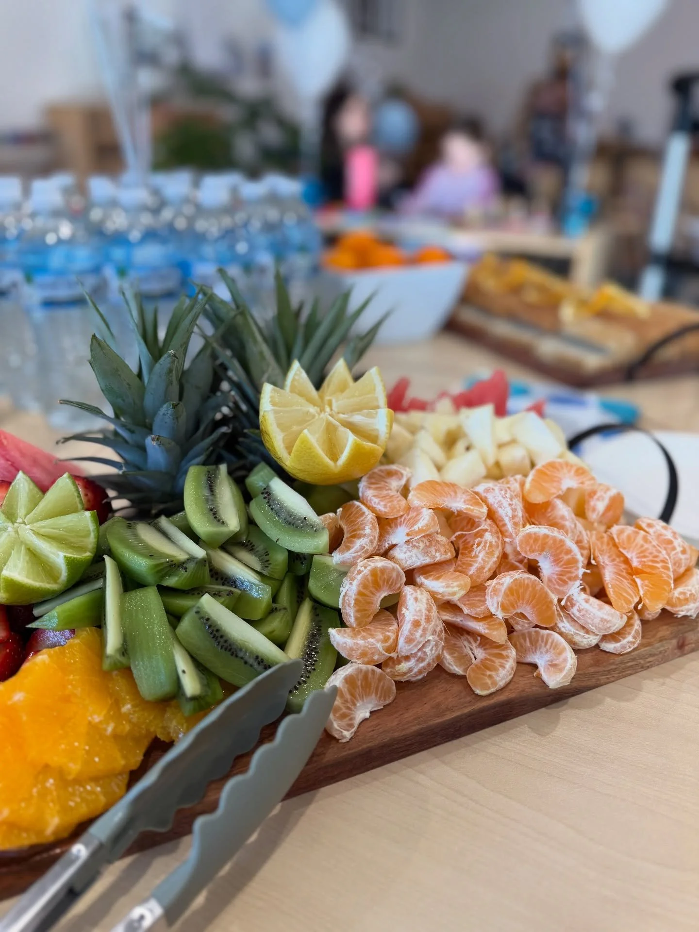 Close-up of a wooden platter filled with sliced fruits including pineapple, lemon, kiwi, mandarin oranges, watermelon, and cantaloupe, with bottled water and blurred people in the background.