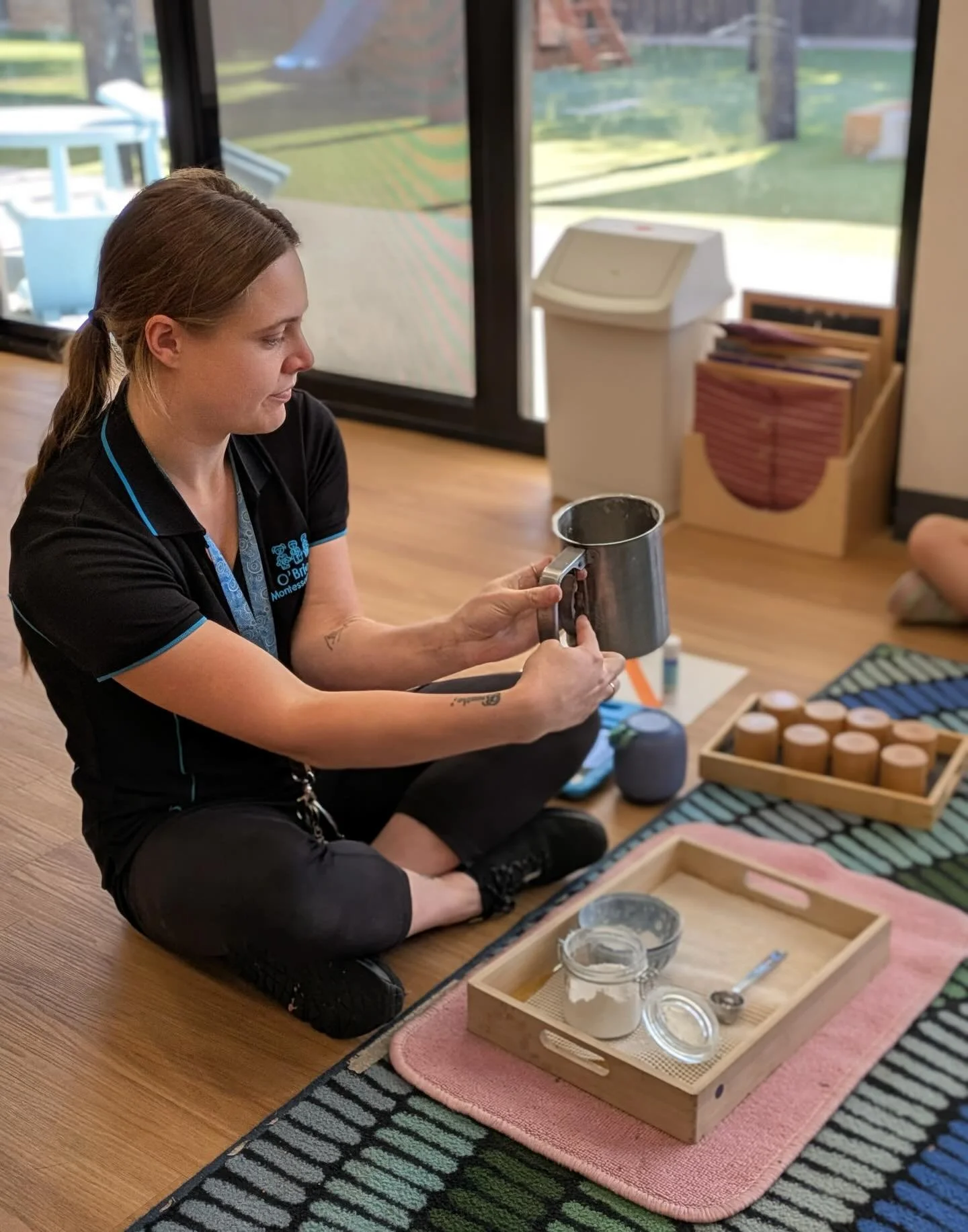 A woman sitting on the floor, engaged in a craft activity with small jars, wooden trays, and craft supplies around her, in a room with large windows and a view outside.
