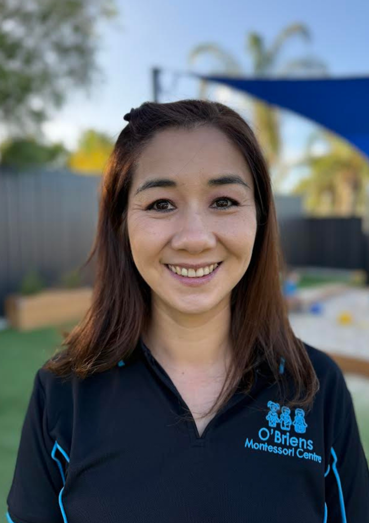 Smiling woman wearing a O'Brien's Montessori Centre shirt outdoors with trees and playground in the background.
