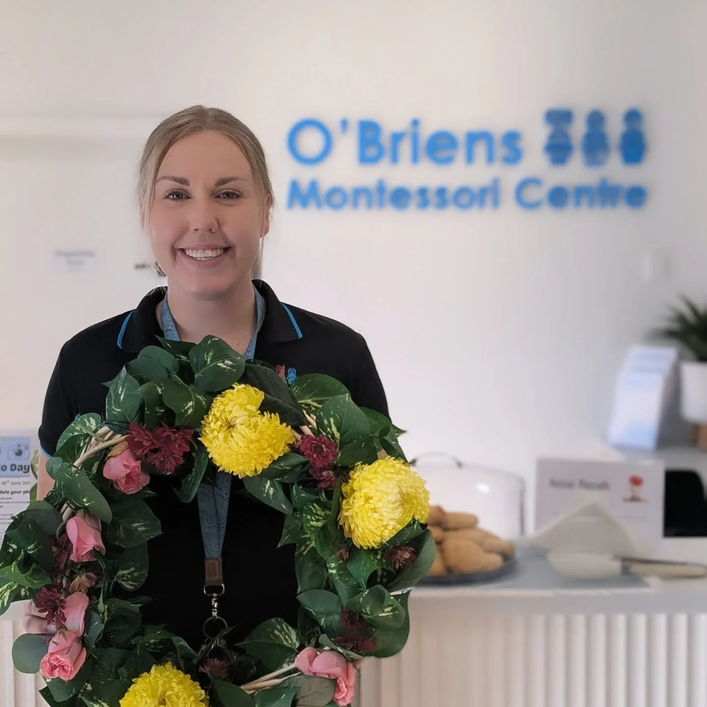 A smiling woman holding a colorful bouquet of flowers in front of a reception desk with blurred signs and items in the background at O'Brien Moncton Centre.
