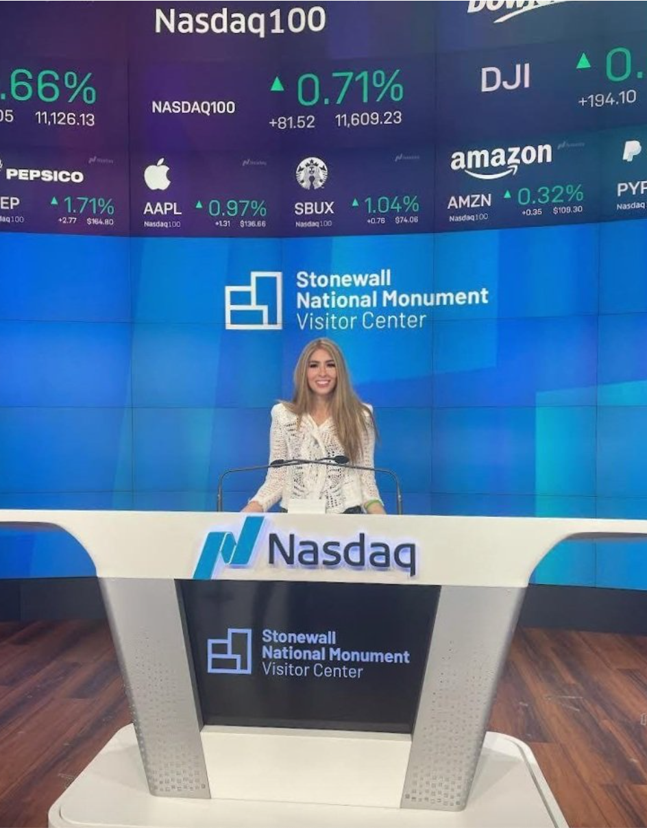 Lisa Winning is smiling and standing at the Nasdaq opening bell. Lisa Winning s in front of a blue background bearing the Stonewall National Monument Visitor Center sign, and Nasdaq stocks.