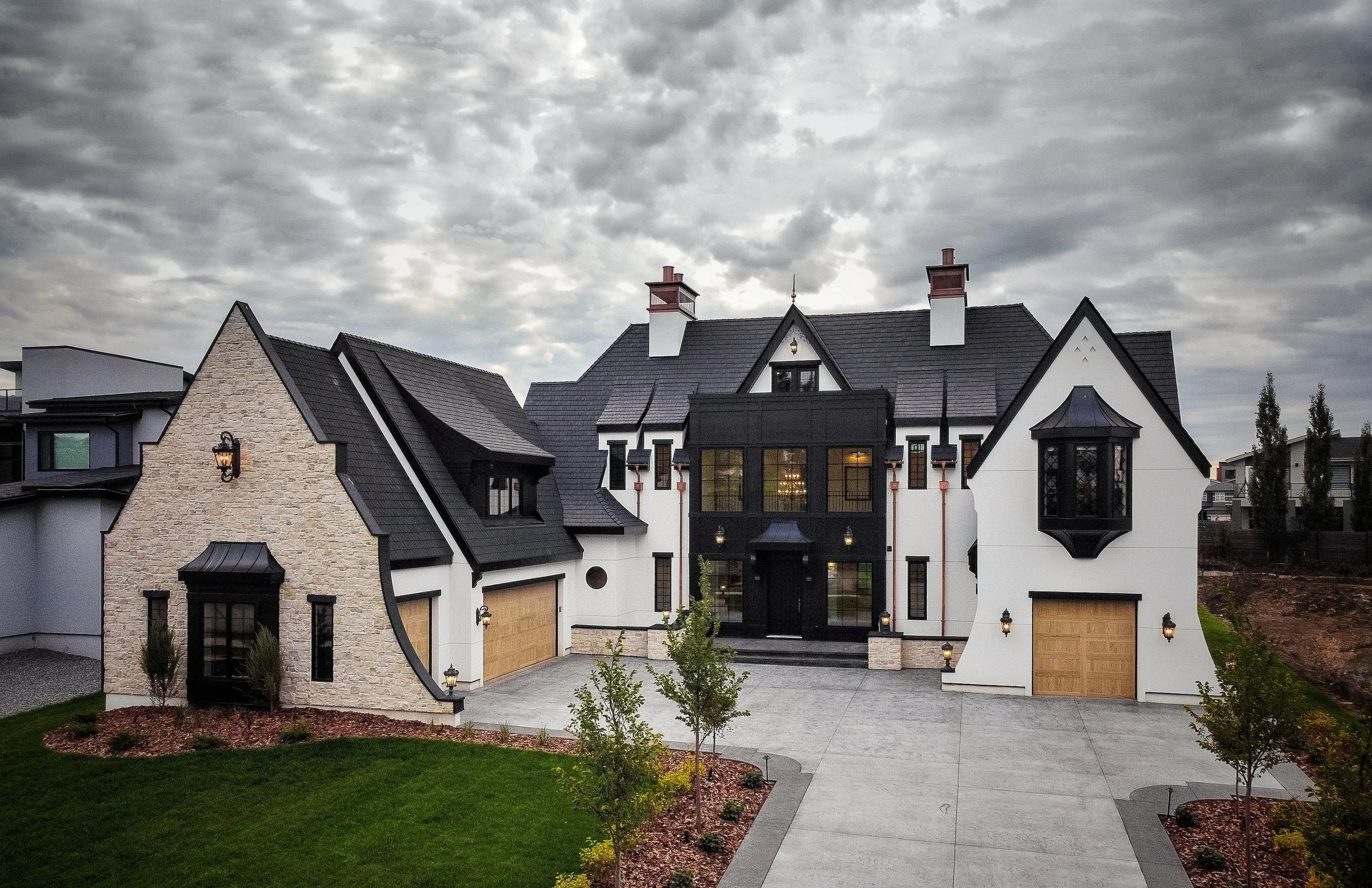 A large, modern Tudor-style house with a mix of stone and white exterior, black roofing, multiple gables, and large windows. The front yard includes a lawn and small trees, with a wide driveway leading to two wooden garage doors.