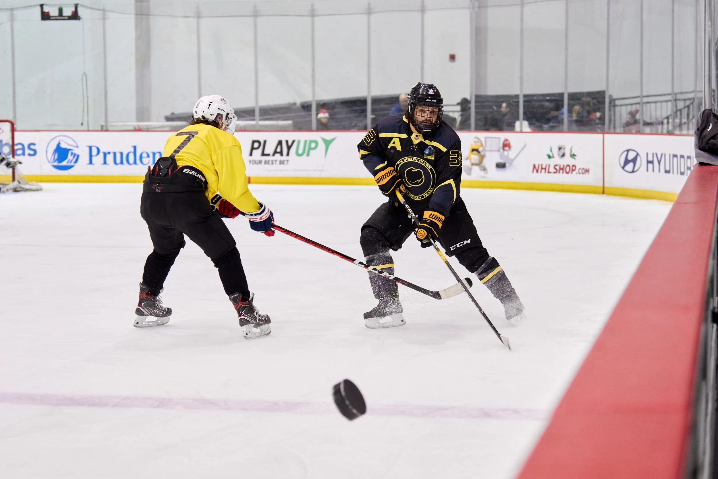 Two ice hockey players in action on an ice rink, one in yellow and the other in dark blue and black, both wearing helmets. The player in yellow is facing the player in dark blue who is moving towards a puck. Advertisements are visible along the rink boards.