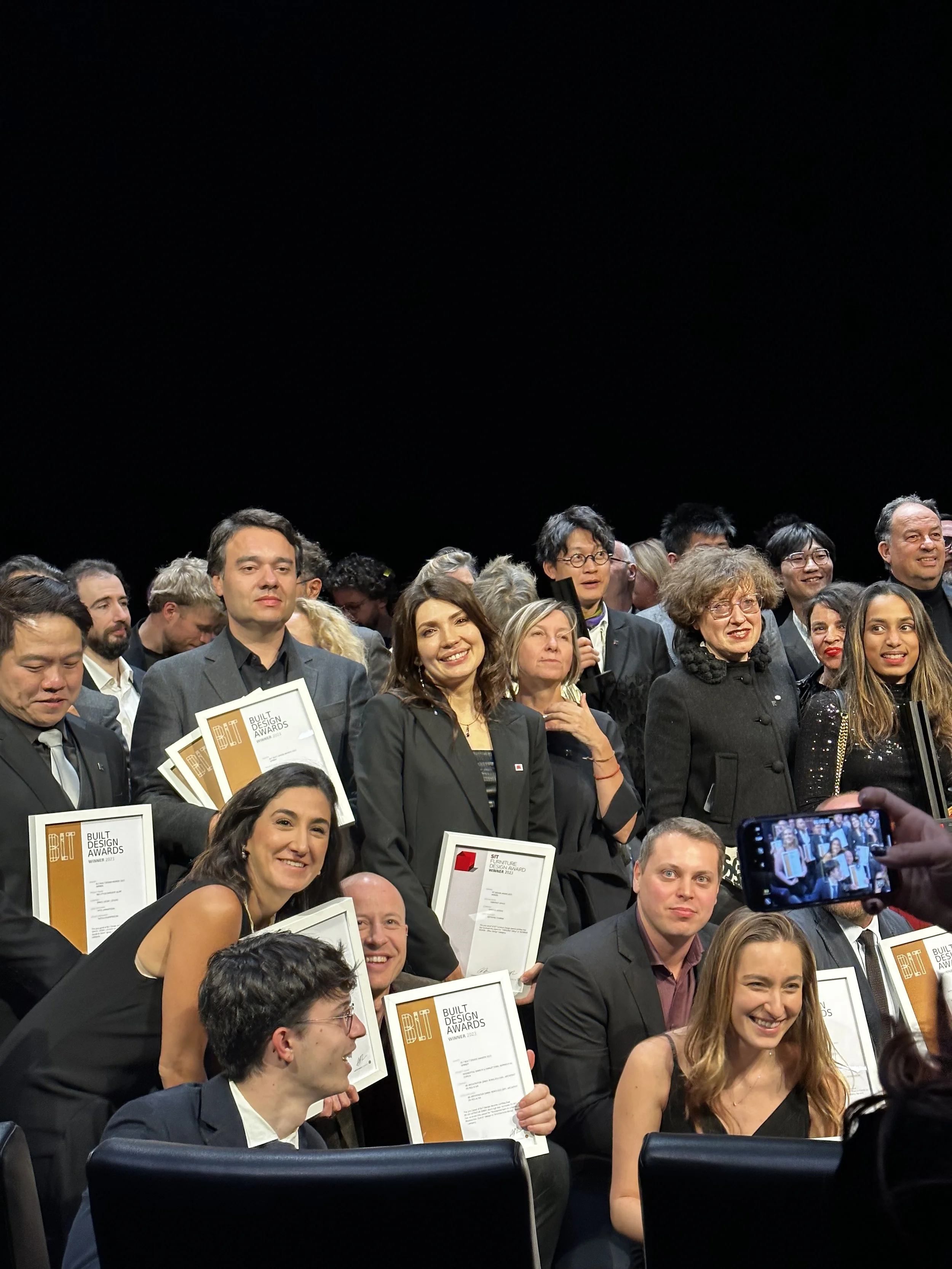 Group of people at an award ceremony holding certificates, smiling for a photo being taken, on a stage with a black background.