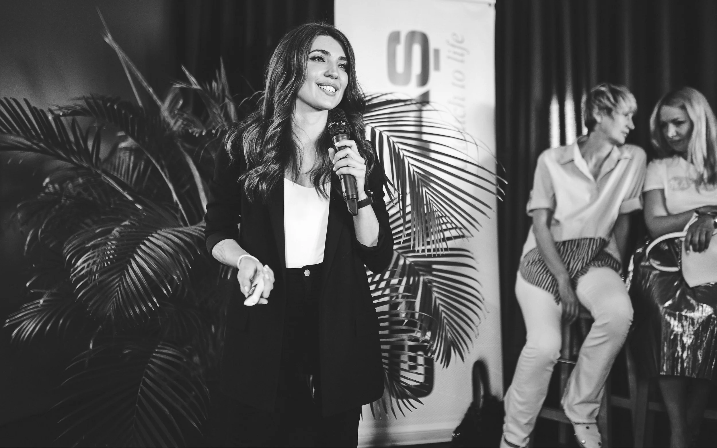 A woman with long wavy hair holding a microphone and smiling, standing in front of a backdrop with large tropical plants behind her, while two women sit and chat in the background.