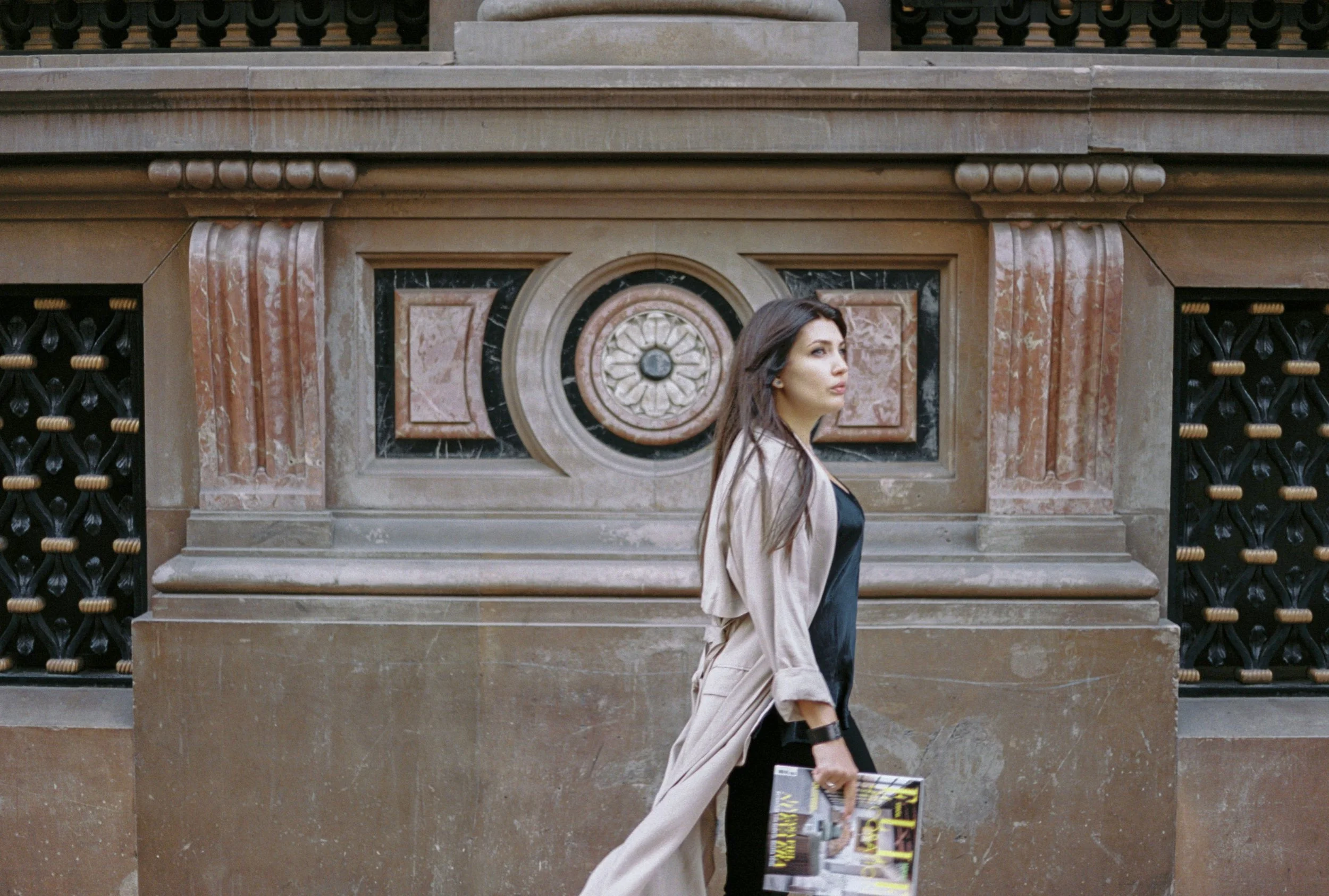 A young woman with long dark hair standing in front of a historic stone building, holding a magazine in her left hand, wearing a beige trench coat over black clothing, looking to the right.