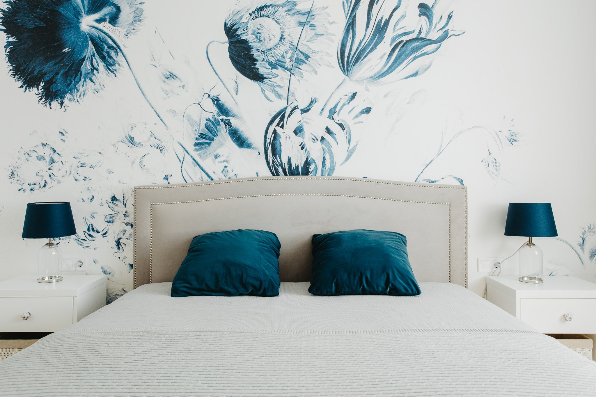 A bedroom with a beige headboard, two blue pillows, white bedding, and matching blue lamps on white nightstands. The wall behind has a large floral mural in shades of blue.